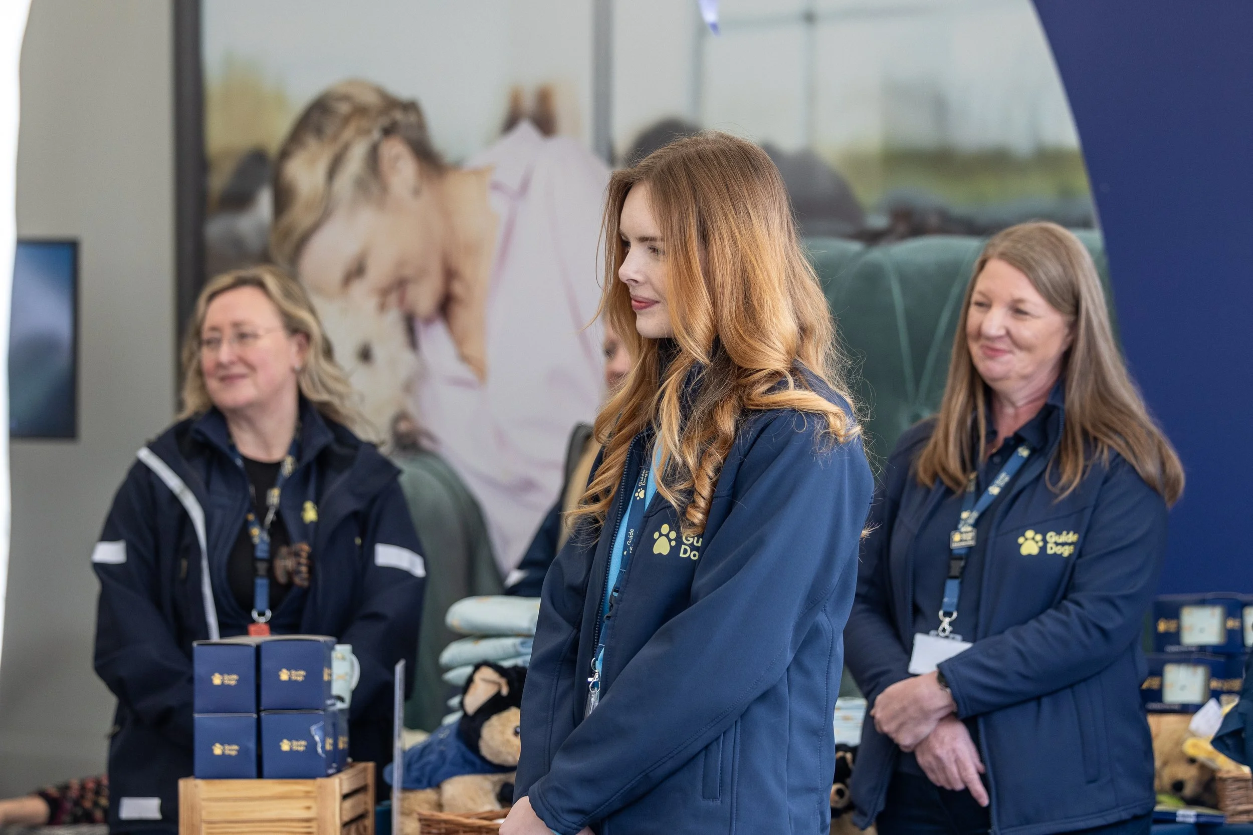 Three women in navy blue jackets with 'Guide Dogs' logos at a booth with pet items and plush toys, indoors with posters in the background.
