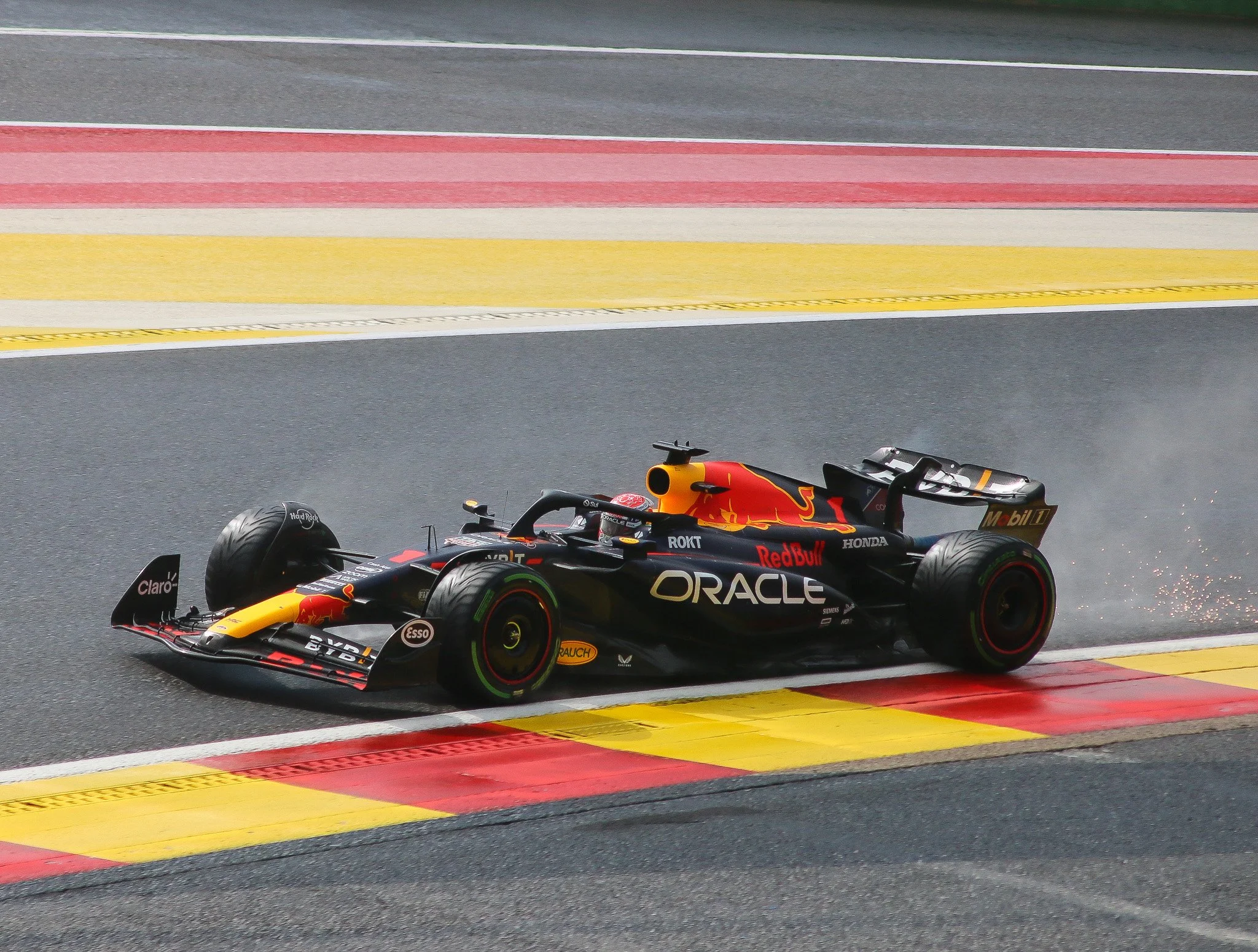 A black Formula 1 race car with Red Bull branding and sponsorship logos, driving on a race track and skidding across the curbing with sparks flying from the underside.