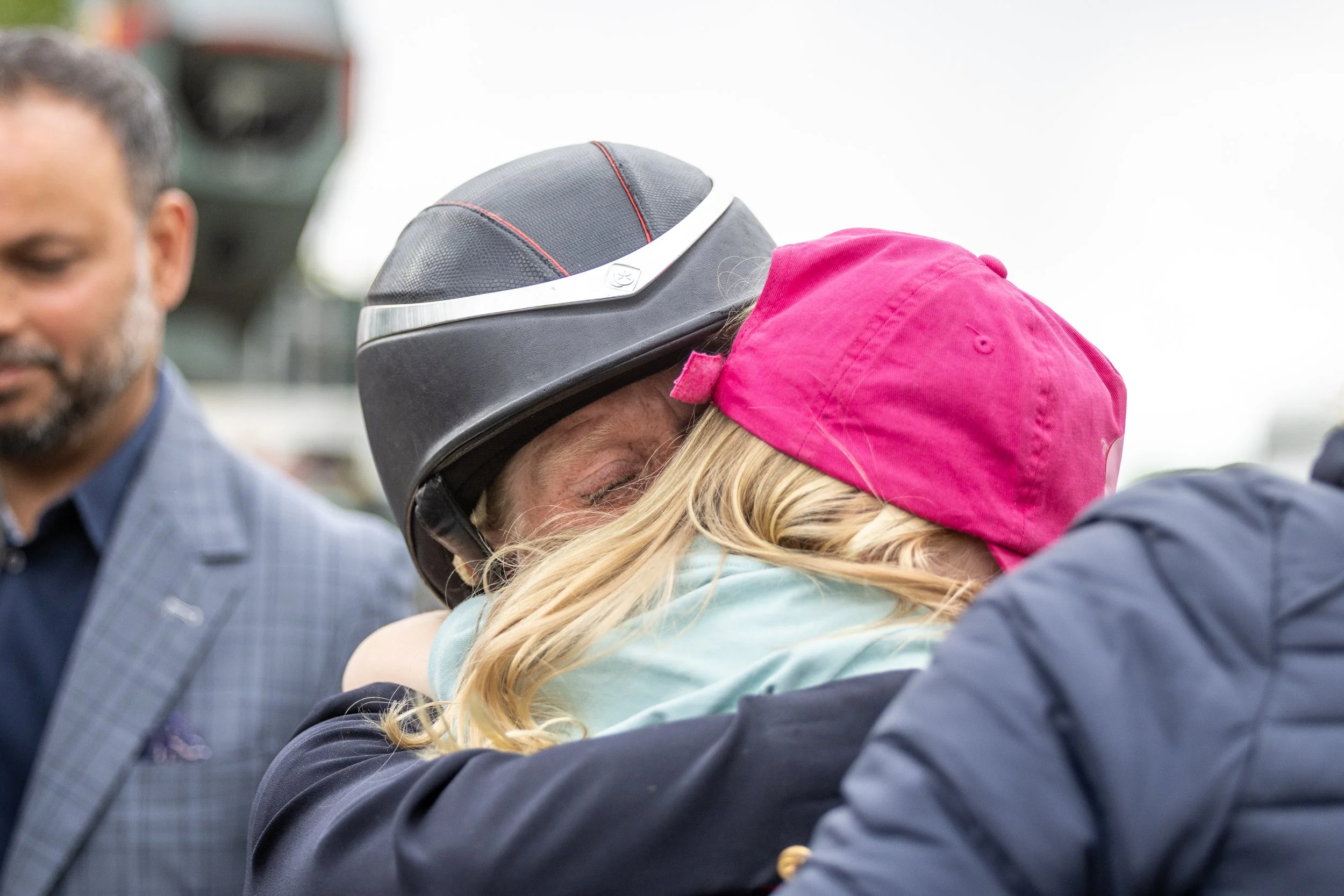 Ros Canter and her daughter, one wearing a black helmet and the other wearing a pink cap, with a man in a blue checkered suit standing nearby. Taken at Badminton Horse Trials after Ros won in 2025.