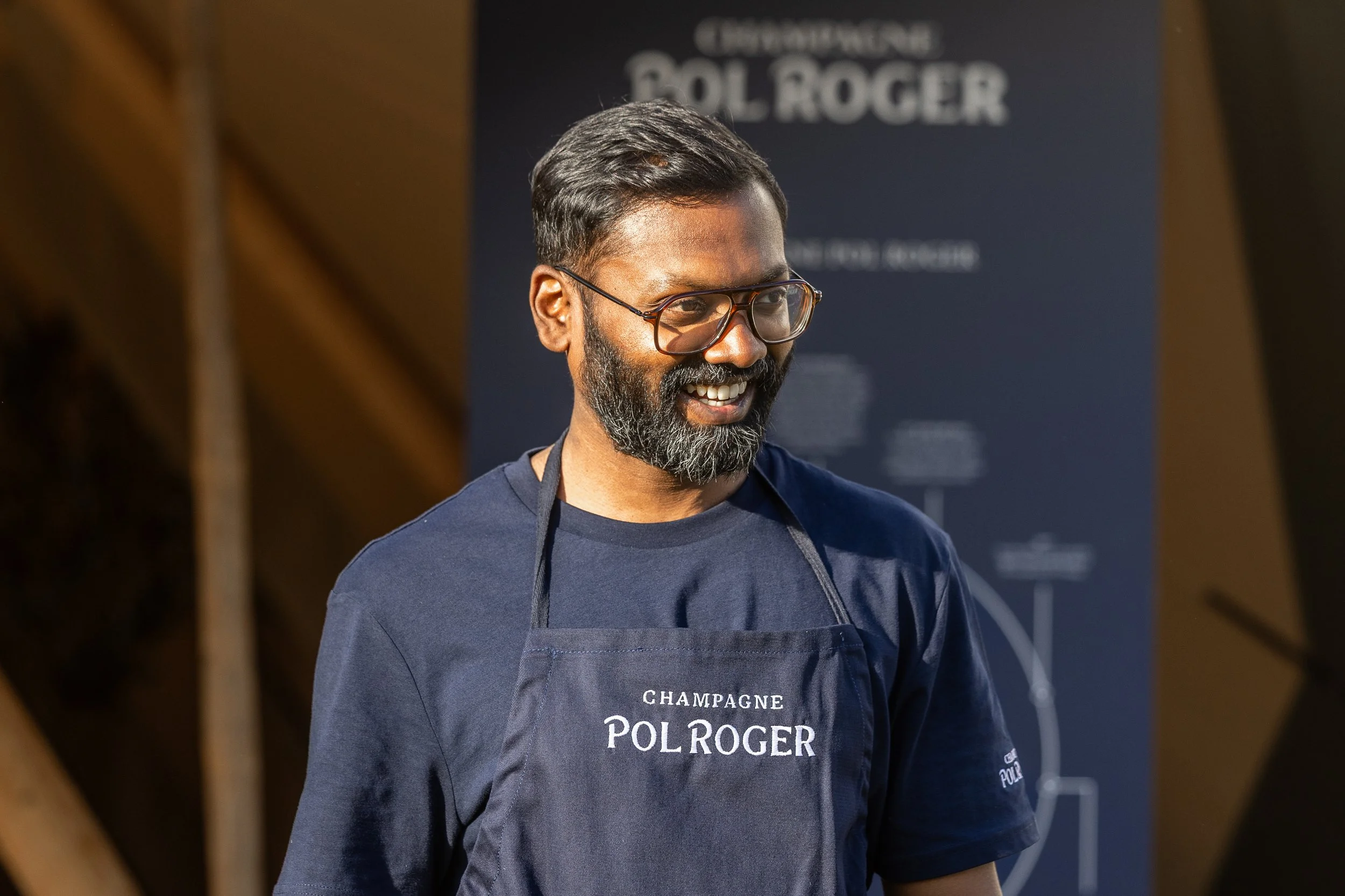 A smiling man with glasses, a beard, and short black hair, wearing a navy blue shirt and apron with "CHAMPAGNE POL ROGER" written on it, standing outdoors in front of a blue sign with white text.