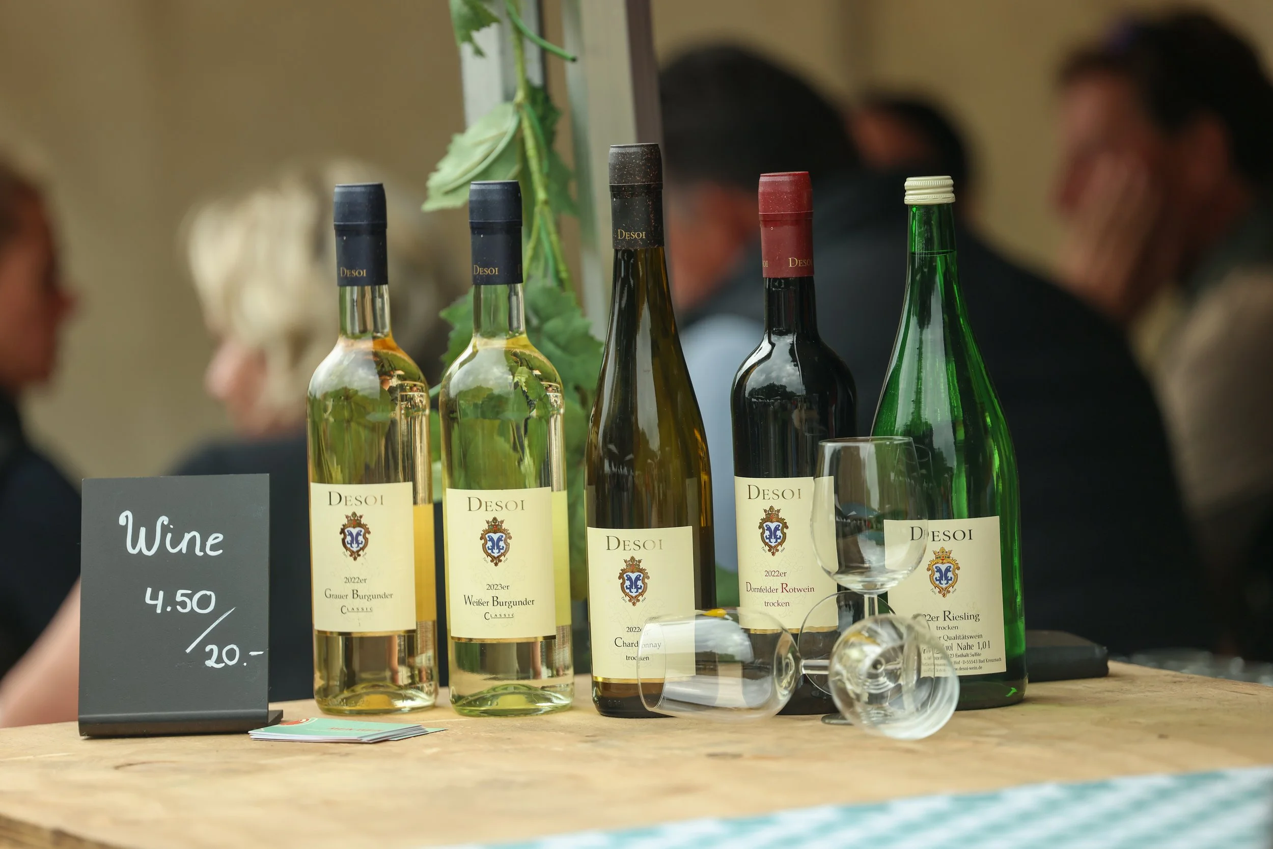 Selection of four bottles of wine on a wooden table, with a small black sign indicating price, and glasses nearby, blurred people in the background.