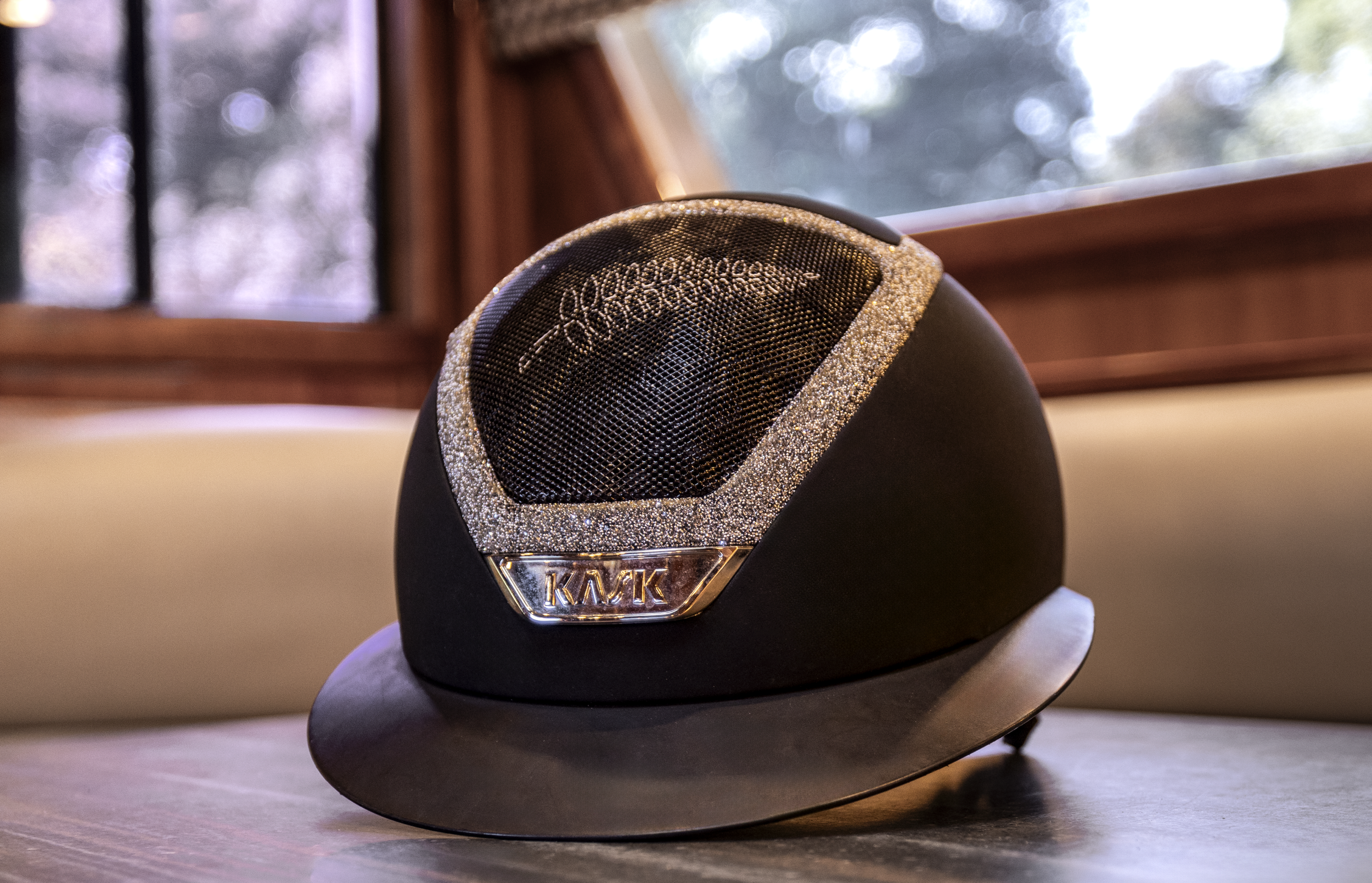 A black 'Kask' helmet with a glittery silver border and a silver badge that says Kask on a wooden surface near a window.