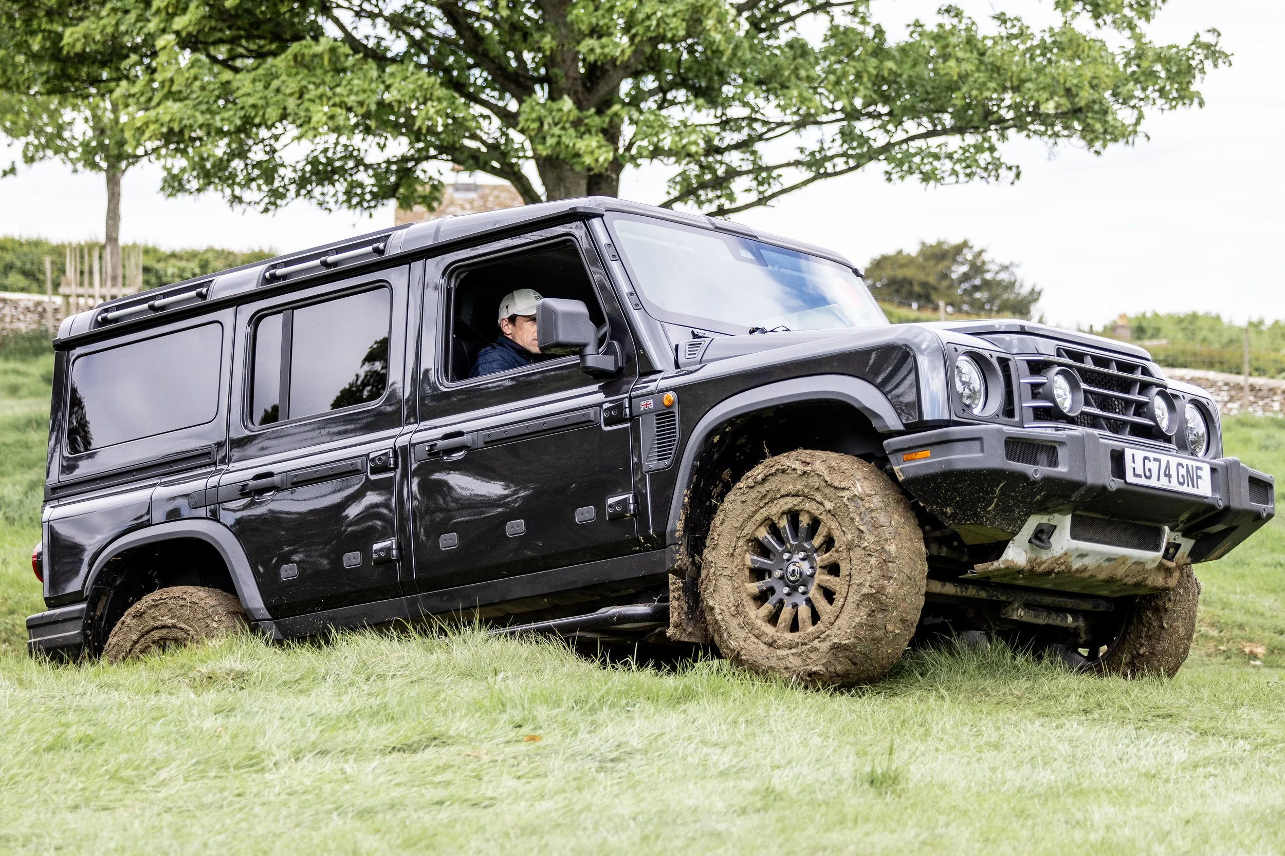 A black 'INEOS' with muddy tires parked on grass under a tree, with a man wearing sunglasses and a cap inside.
