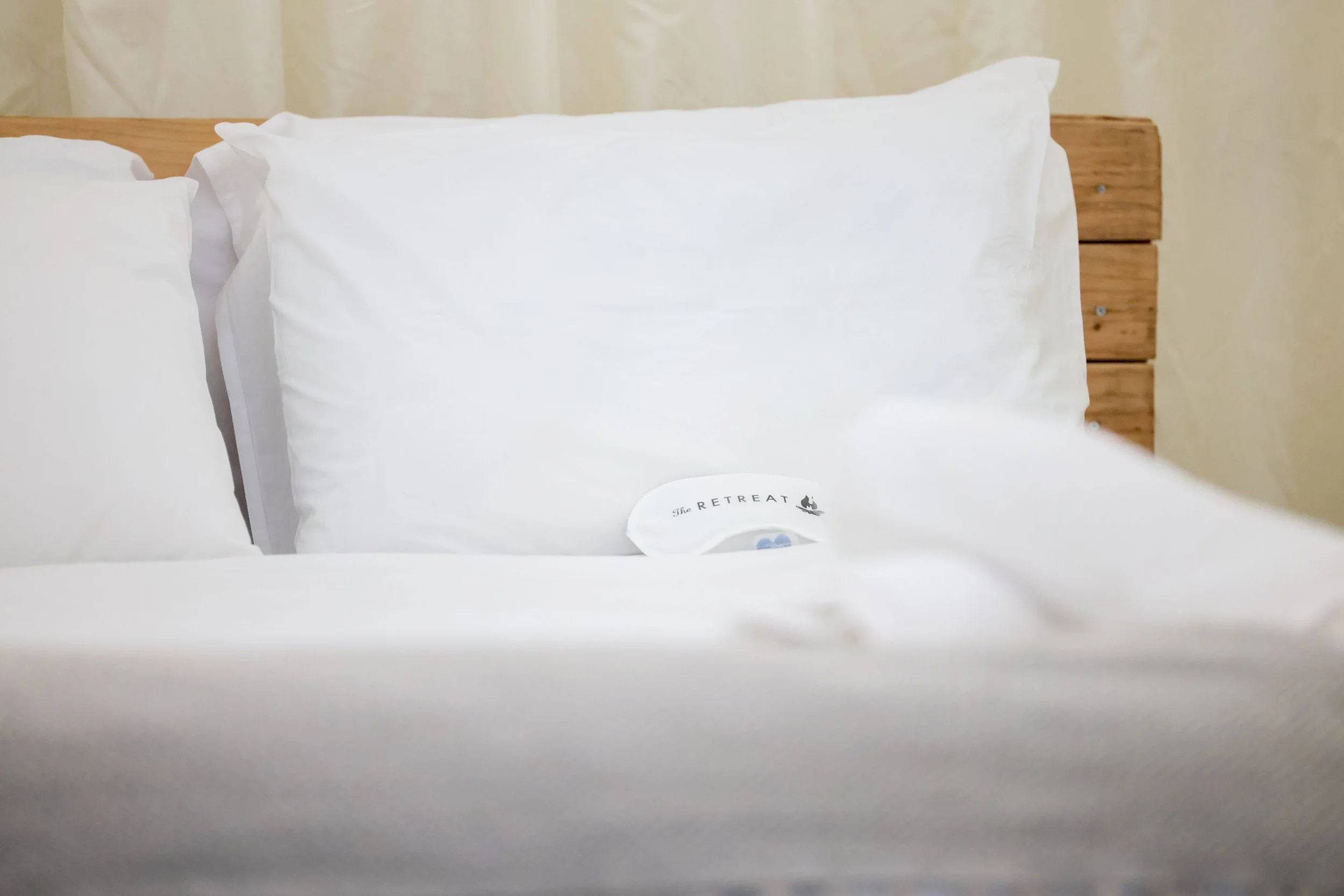 A neatly made bed with white pillows and sheets, featuring a small fan with the label 'The Retreat' placed among the pillows. The headboard is made of wooden planks, and a beige curtain is visible in the background.