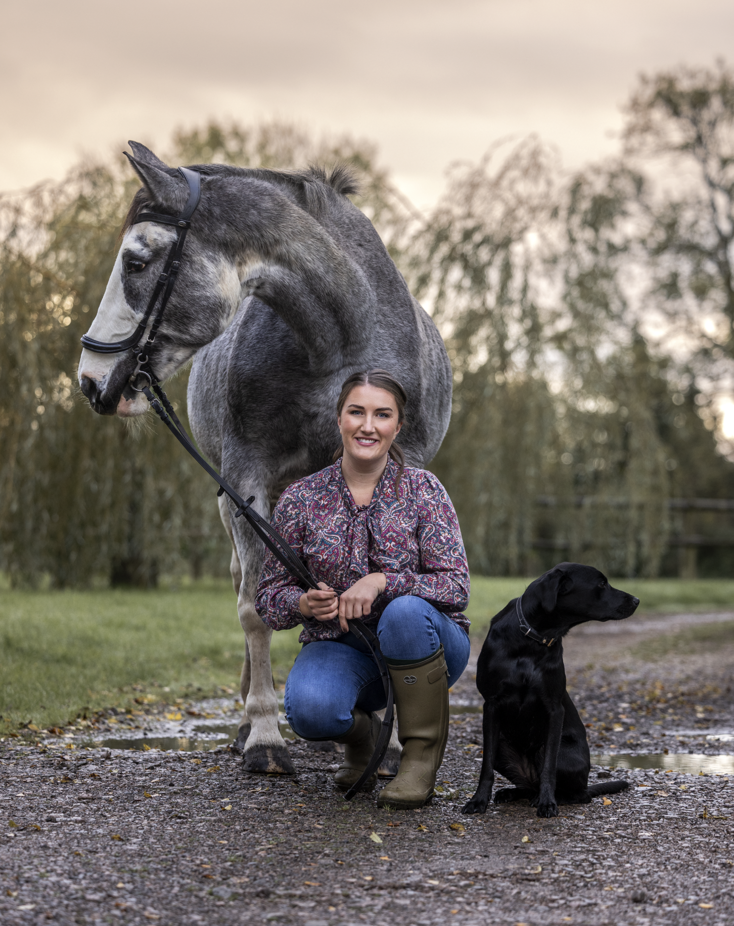 A smiling woman kneeling on a gravel path with a black Labrador retriever sitting beside her and a large gray and white horse standing behind her outdoors during sunset.