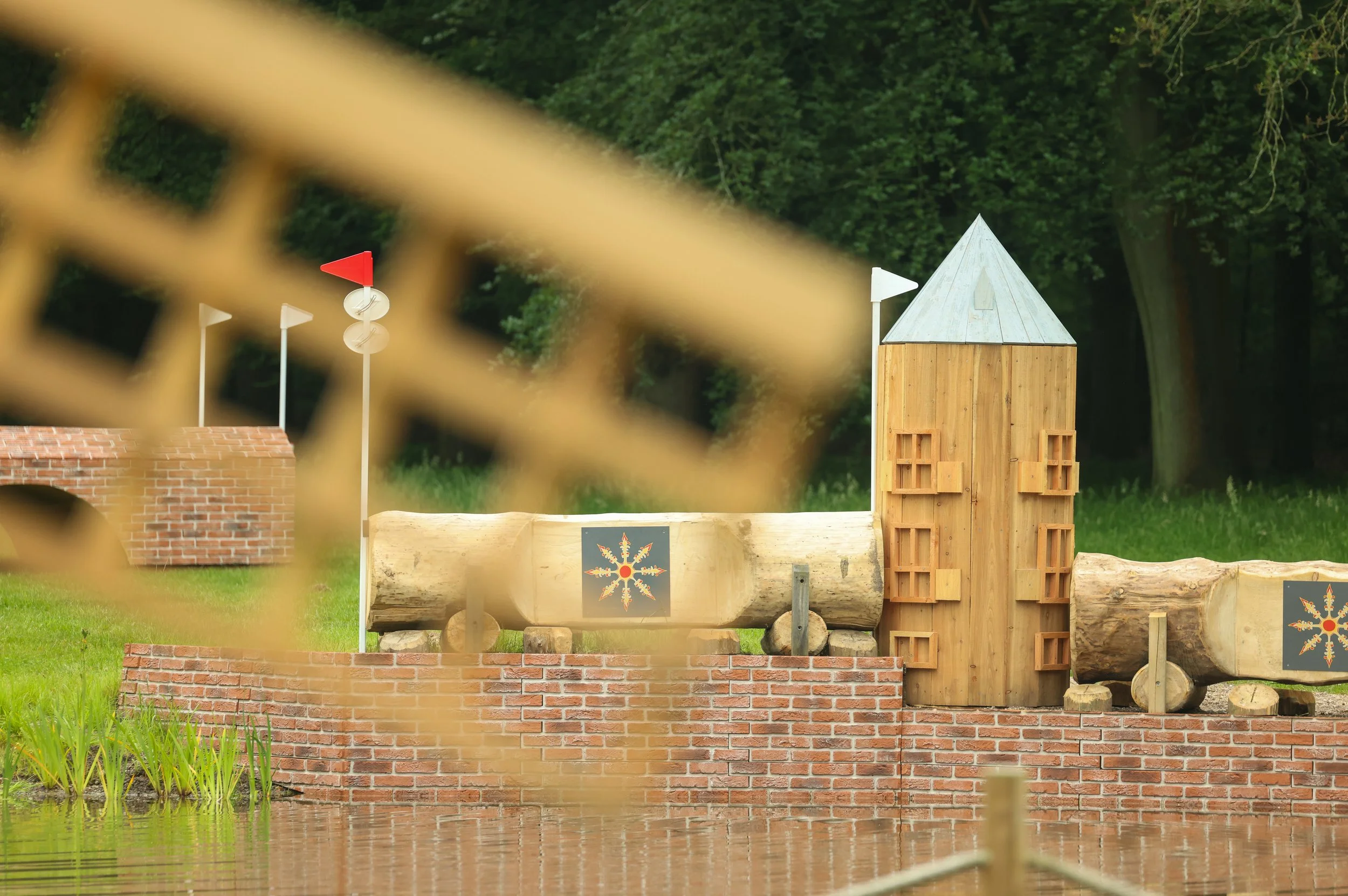 Wooden structure resembling a small tower or building with logs and bricks around it, set in a grassy area with trees in the background, partially viewed through a fence. Taken at Maarsbergen Horse Trials