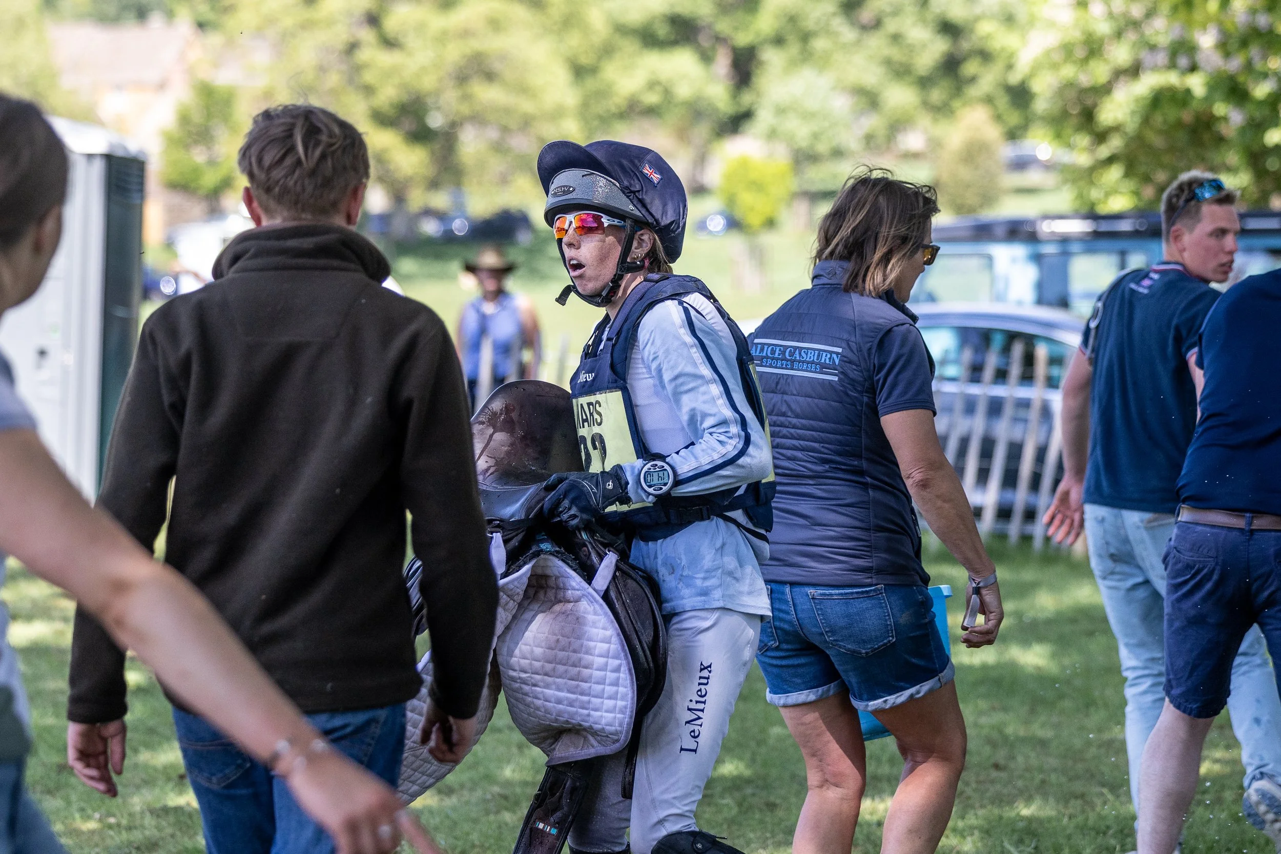 A female after cross country wearing helmet, sunglasses, and white sports gear standing on grass, holding a helmet, surrounded by spectators and event staff.