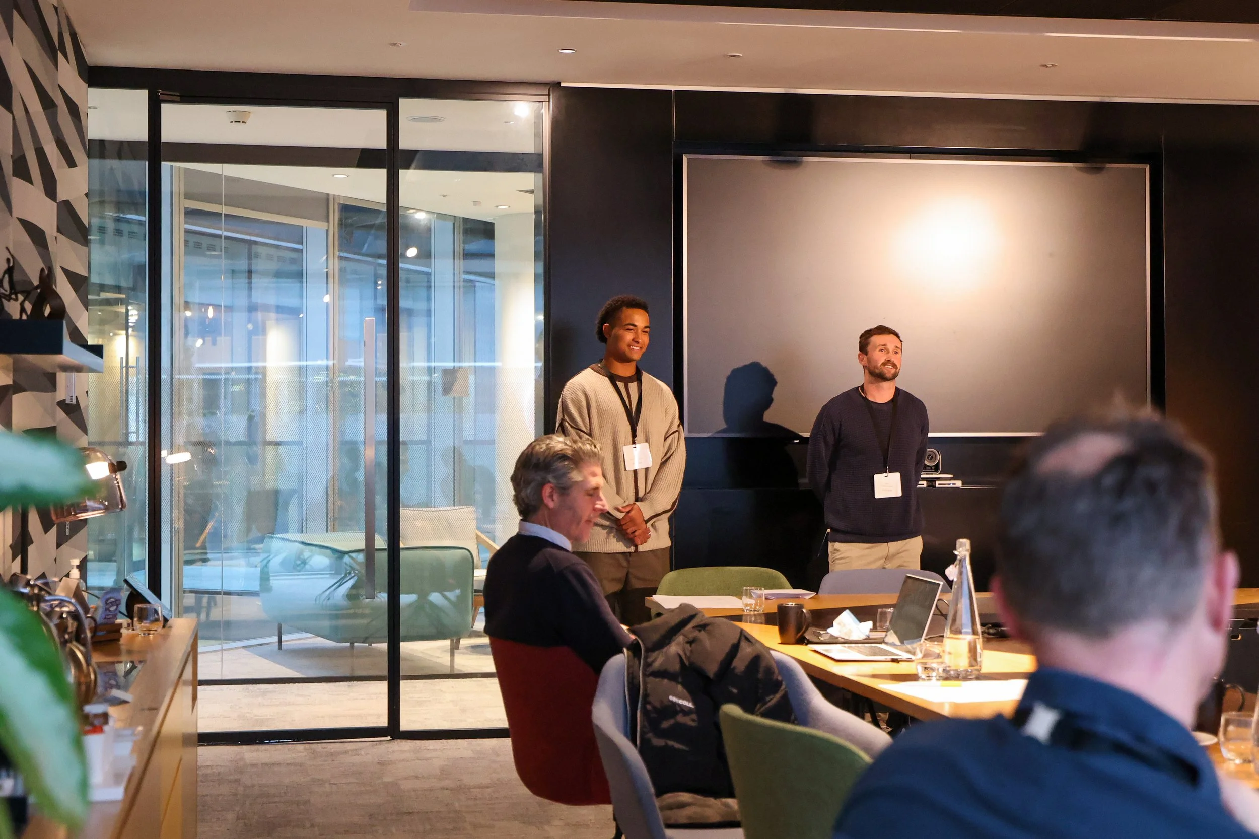 Two men standing at the front of a conference room, speaking to an audience. One man is smiling, wearing a beige sweater, and the other has a beard, wearing a dark sweater. Audience members are seated at the table, some taking notes, others listening