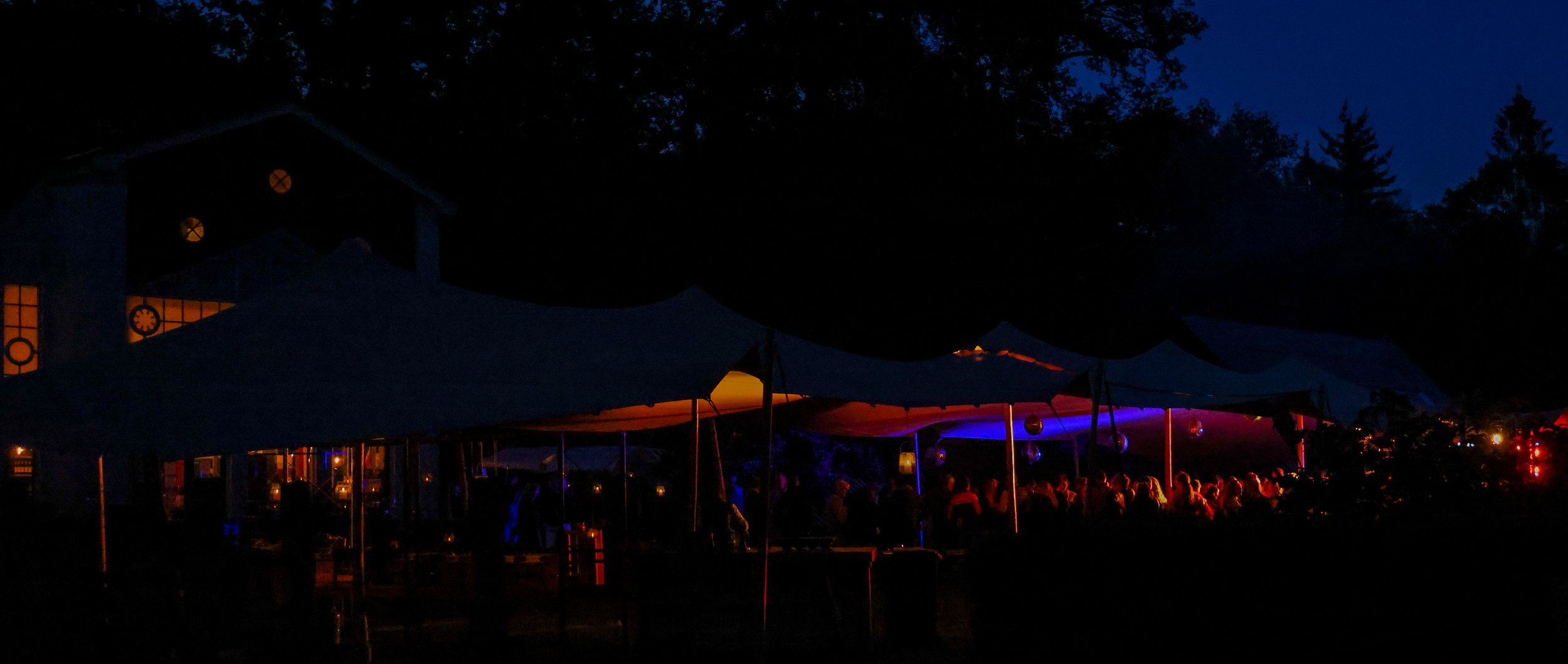 Nighttime outdoor gathering with tents illuminated by colorful lights, and a crowd of people under the canopy.
