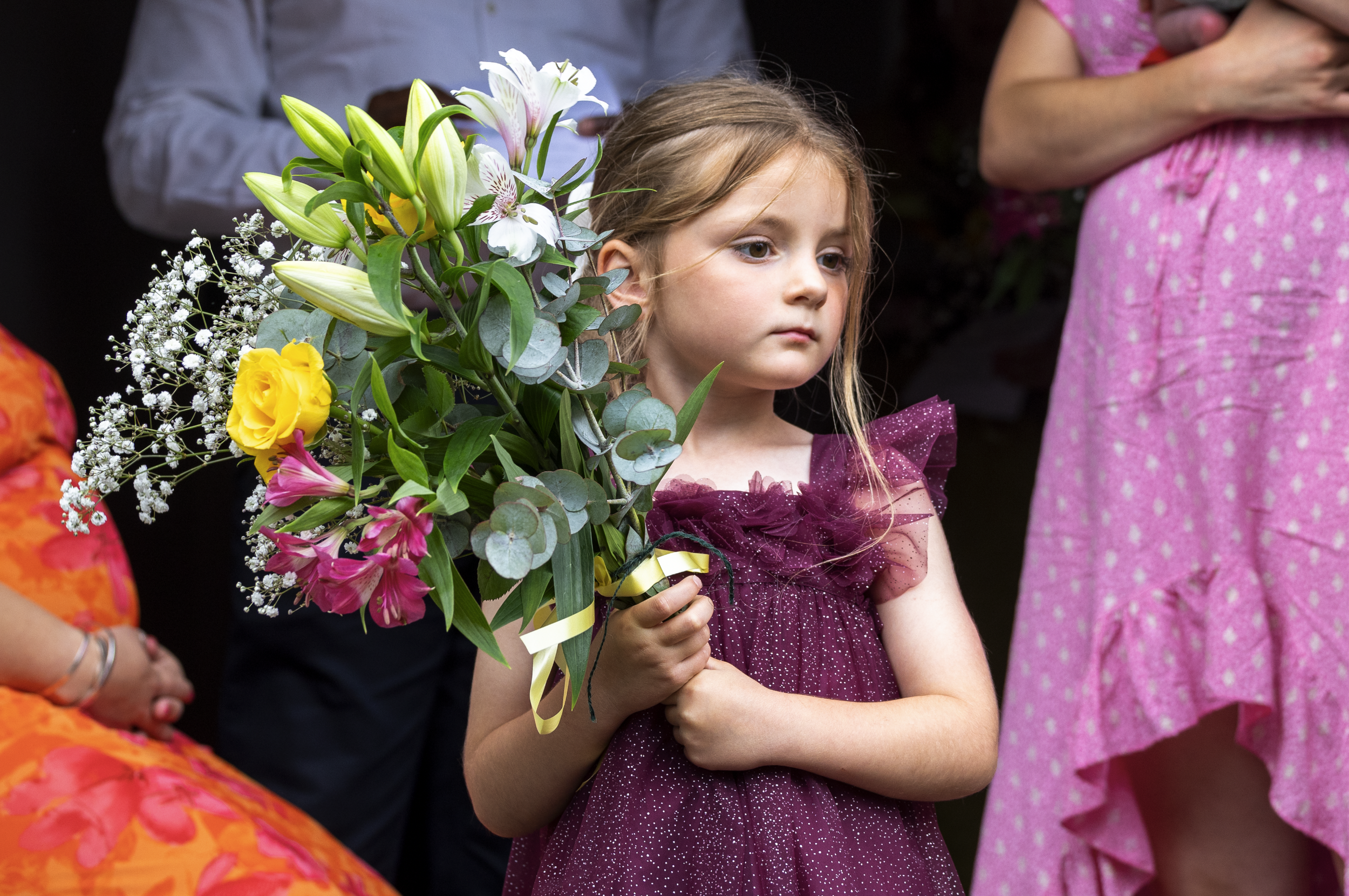A young girl in a purple dress holding a large bouquet of flowers with lilies, roses, and other blooms during a celebration or ceremony.