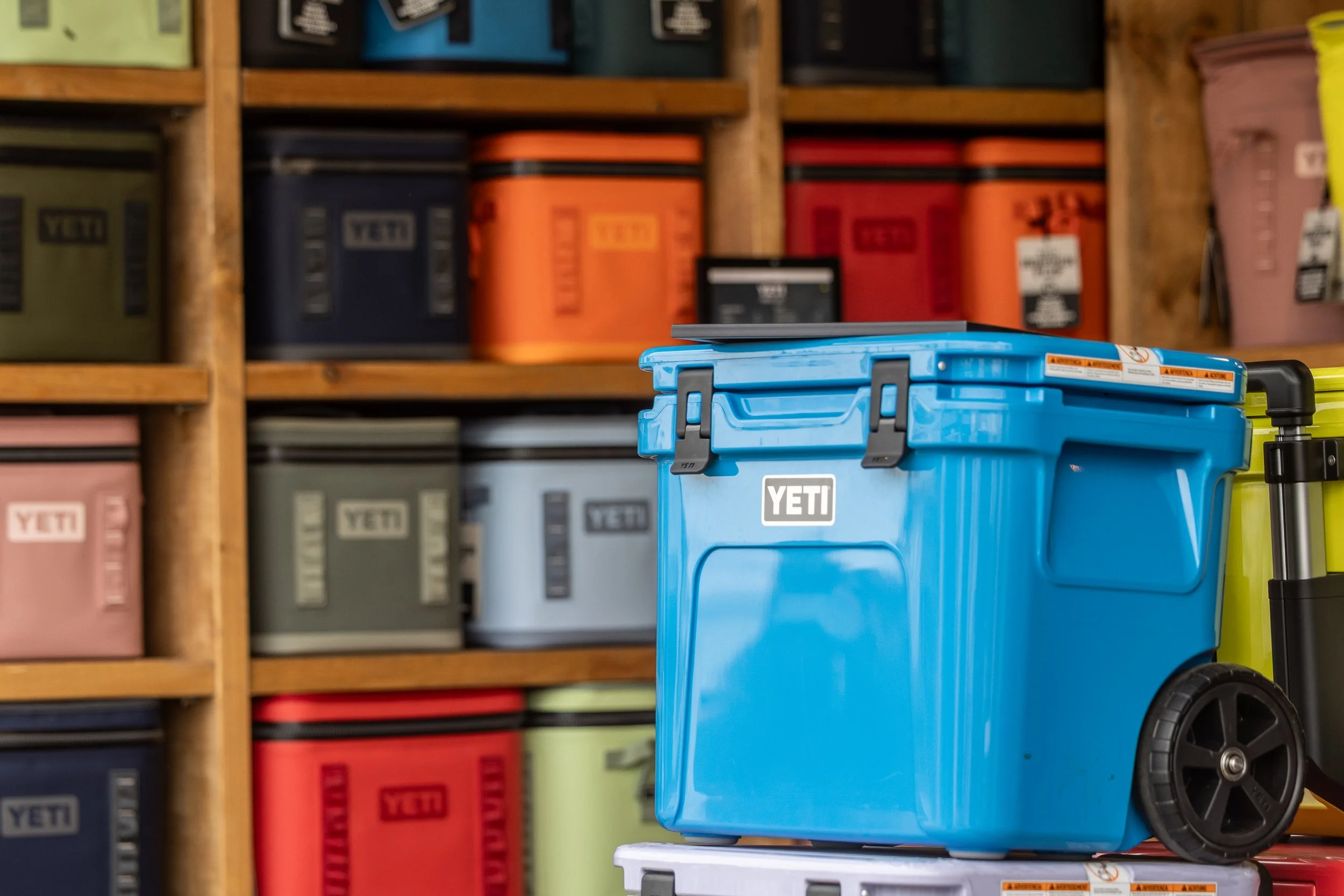 A blue YETI cooler with black wheels and handle in front of a wooden shelf filled with various colored YETI coolers.