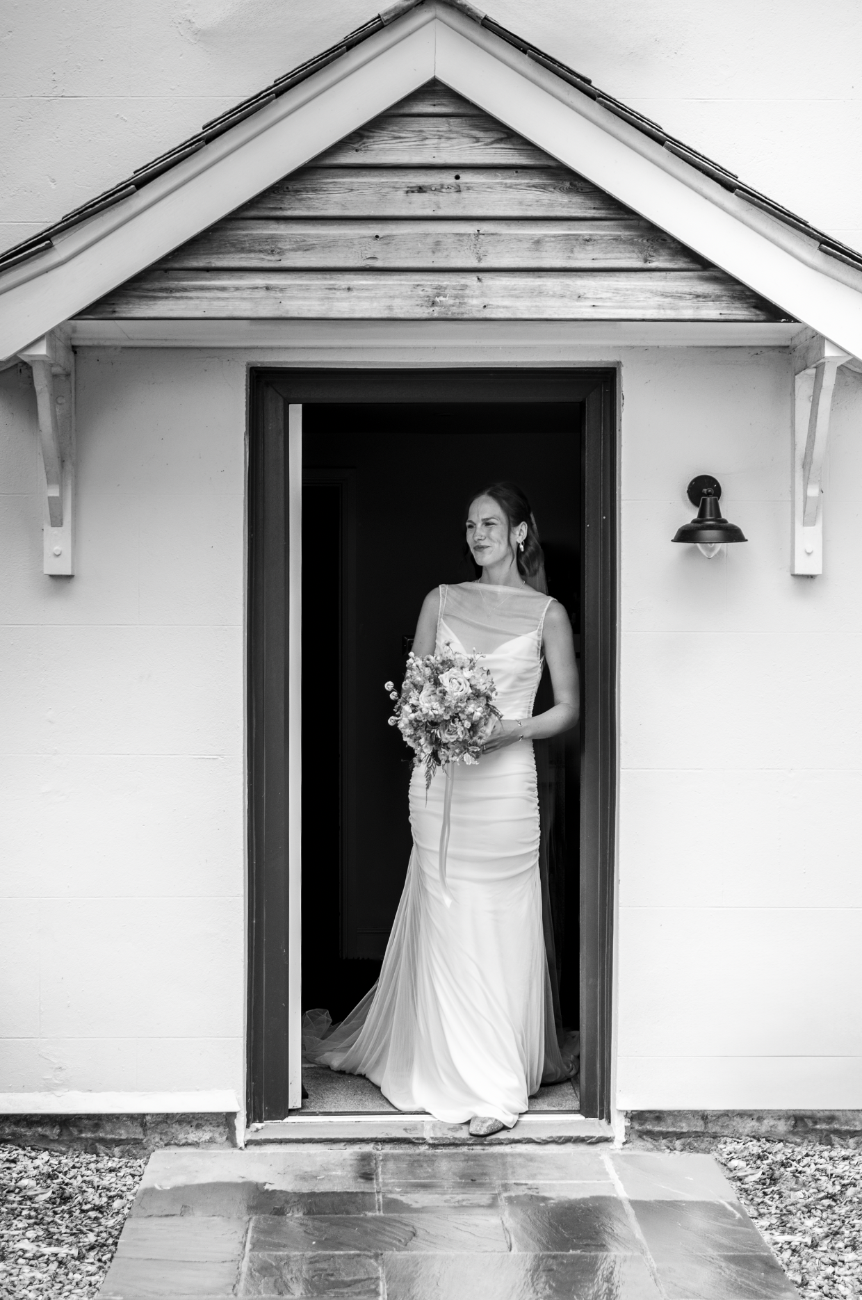 Black and white photo of a bride standing in a doorway, holding a bouquet of flowers, wearing a wedding gown, looking to the side with a smile.