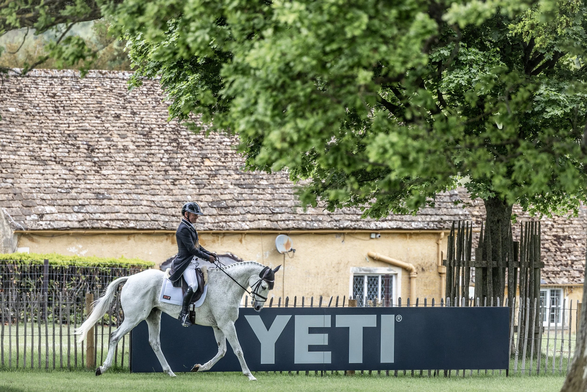 A person riding a white horse in an outdoor setting with a large green tree and an old yellow building in the background, near a YETI sign and a wooden fence.