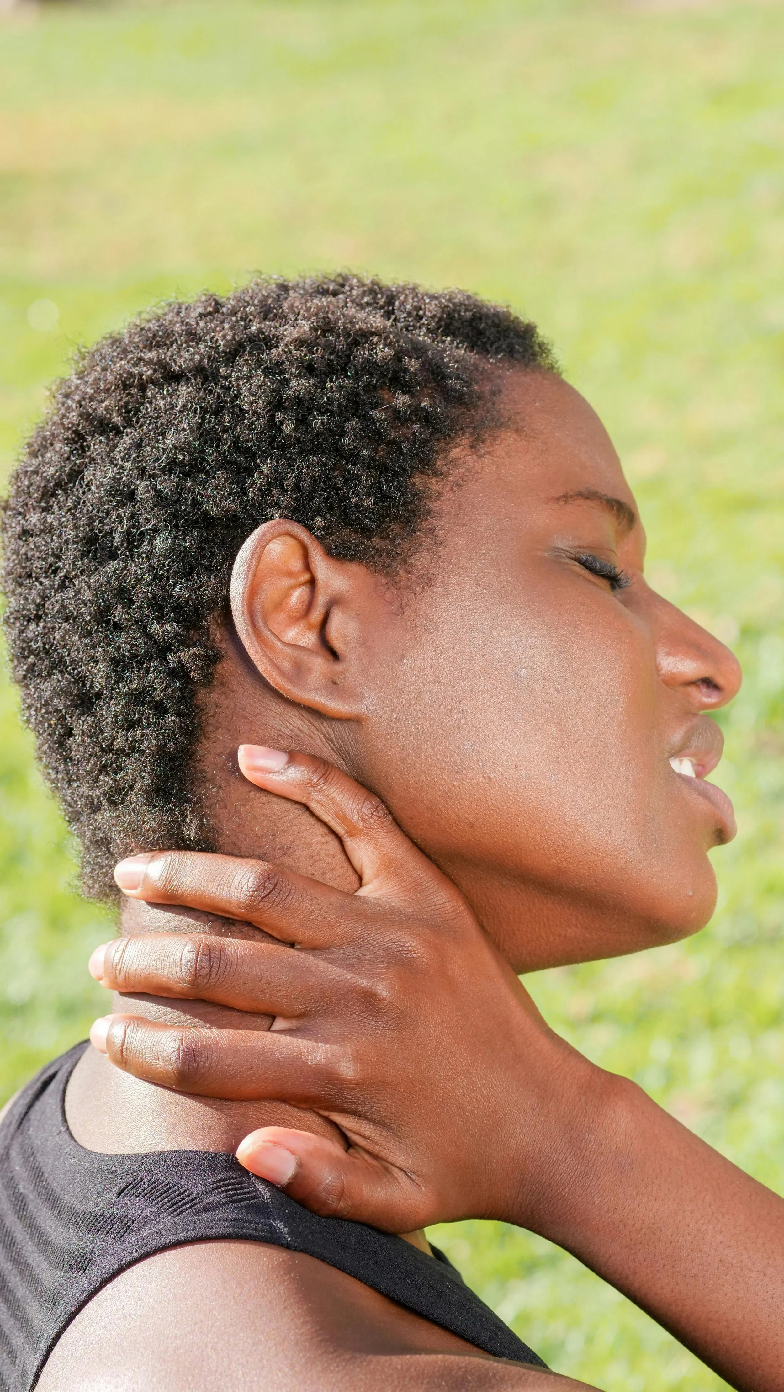 A woman with short, curly black hair is holding her neck with her right hand and appears to be experiencing discomfort or pain. She is outdoors, with a blurred green background, and is wearing a black sleeveless top.