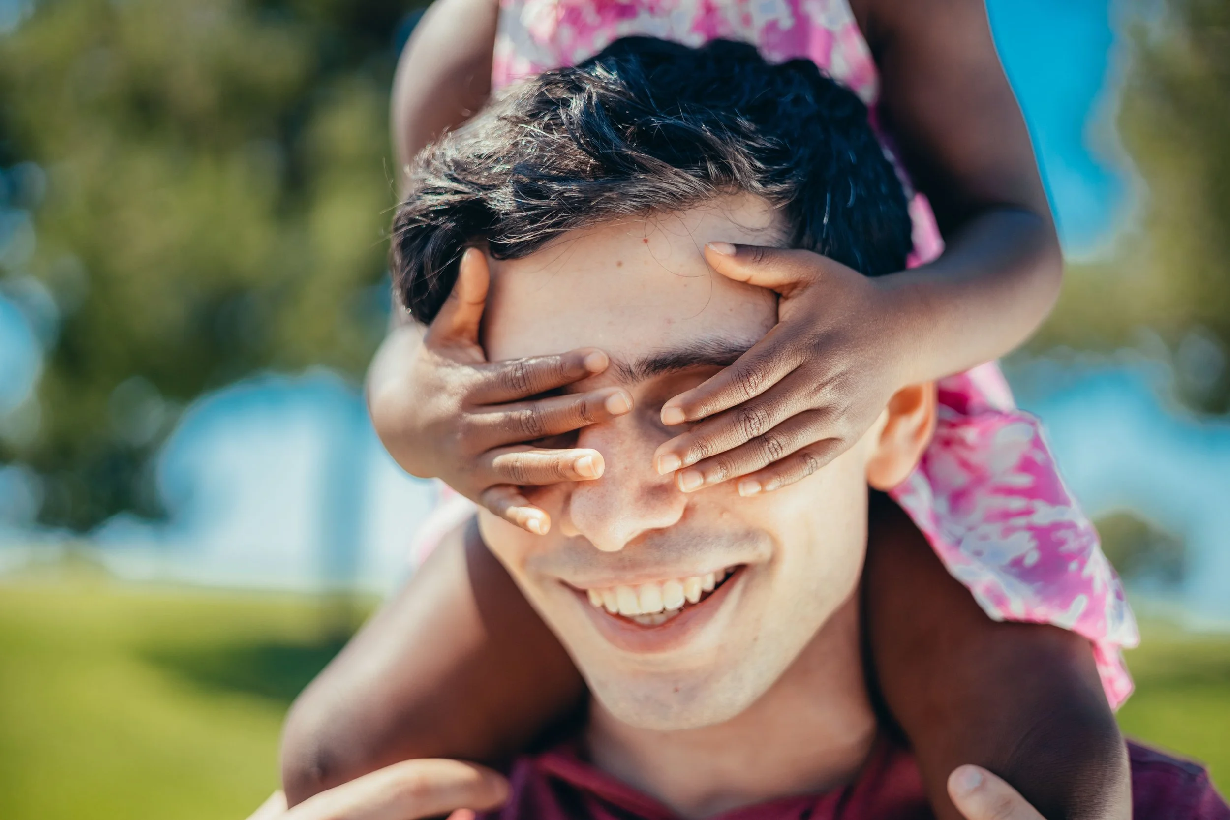 “Smiling father carrying a child on his shoulders outdoors as the child playfully covers his eyes.”