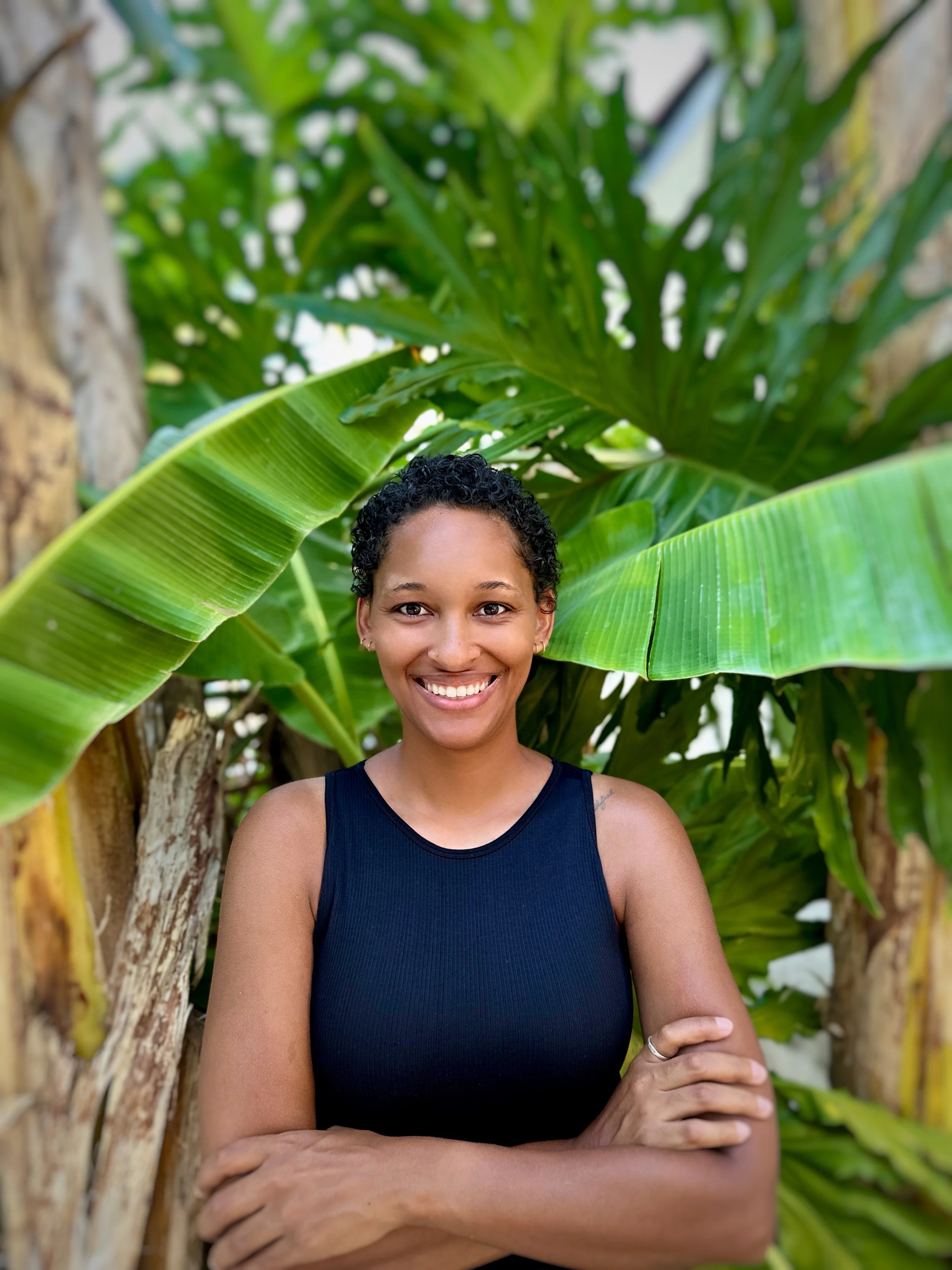 A woman smiling outdoors with large green tropical leaves in the background.