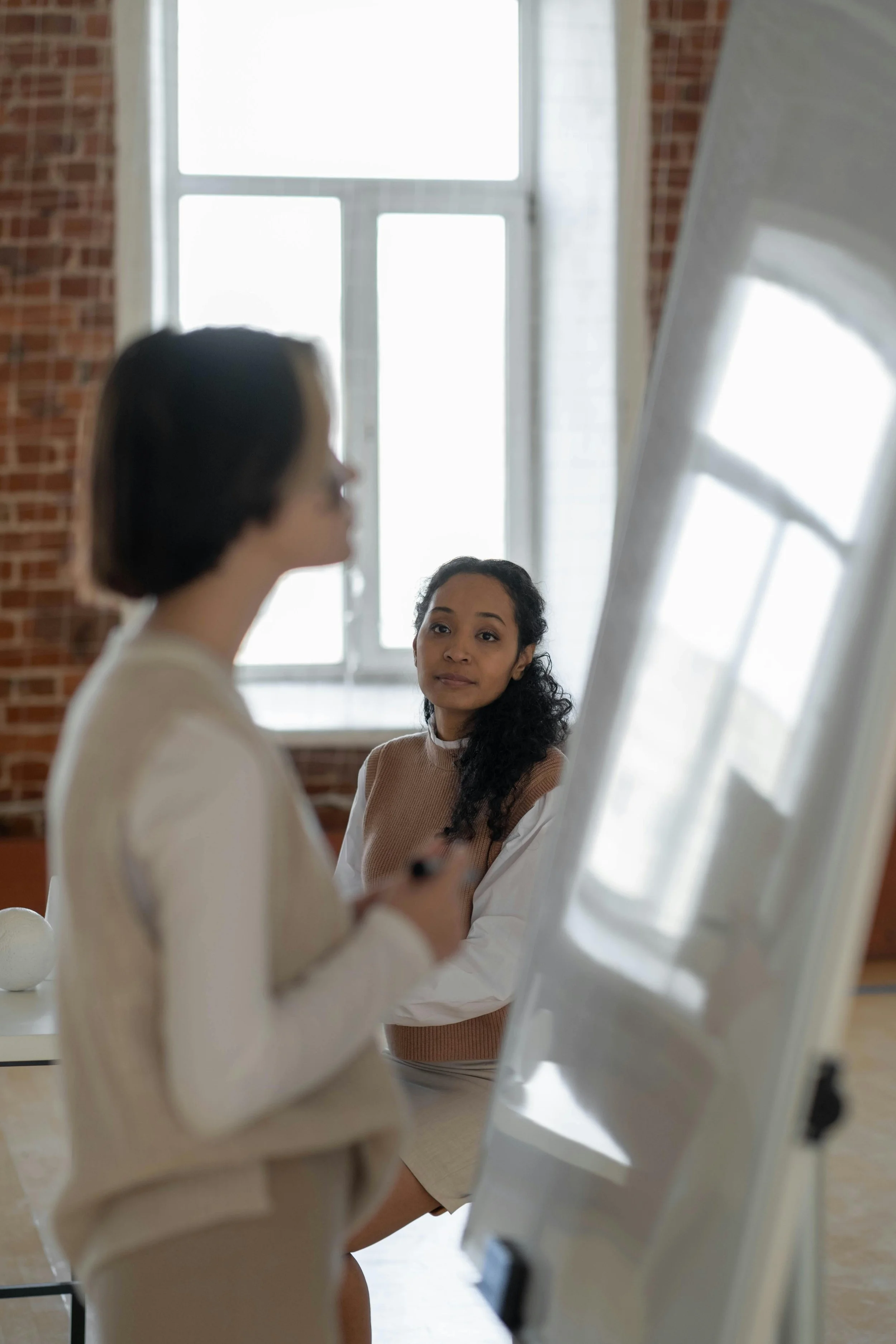 A woman with curly black hair sits in front of a large window, looking at another woman who is standing and speaking in front of a whiteboard or flip chart in a bright room with brick walls.