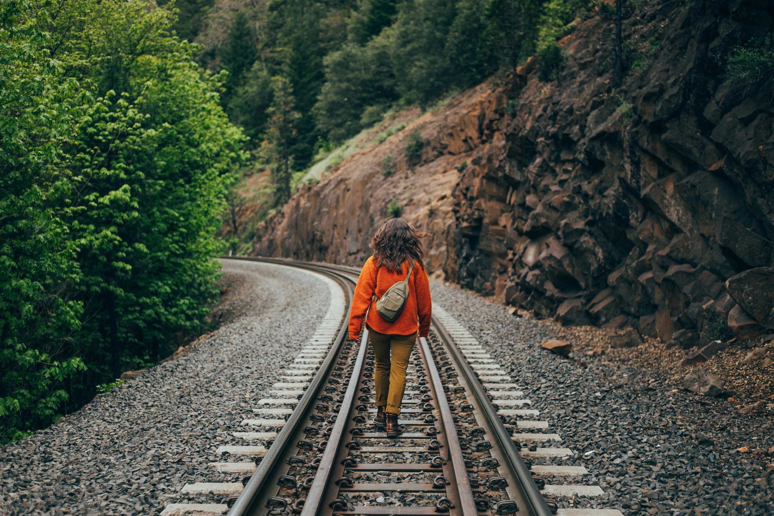 A woman walking along a railway track through a lush, green forested area with rocky cliffs on one side.