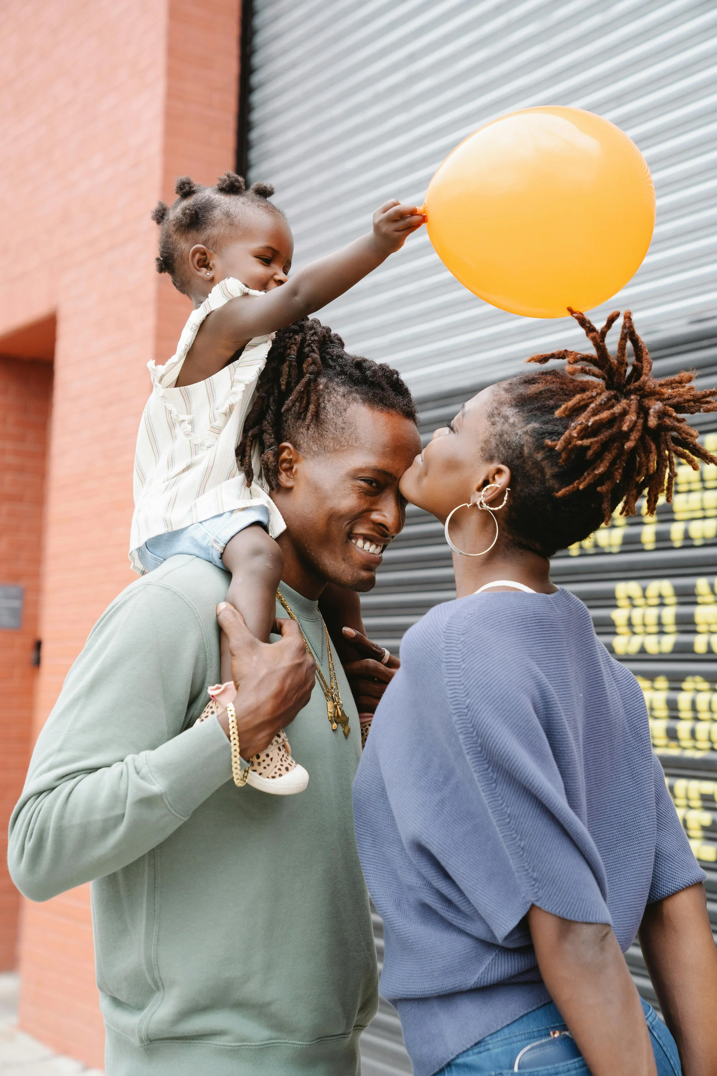 A happy family of three outdoors near a brick building with a roller shutter. The father is carrying a young girl on his shoulders who is holding a yellow balloon, and a woman is kissing the father's cheek.