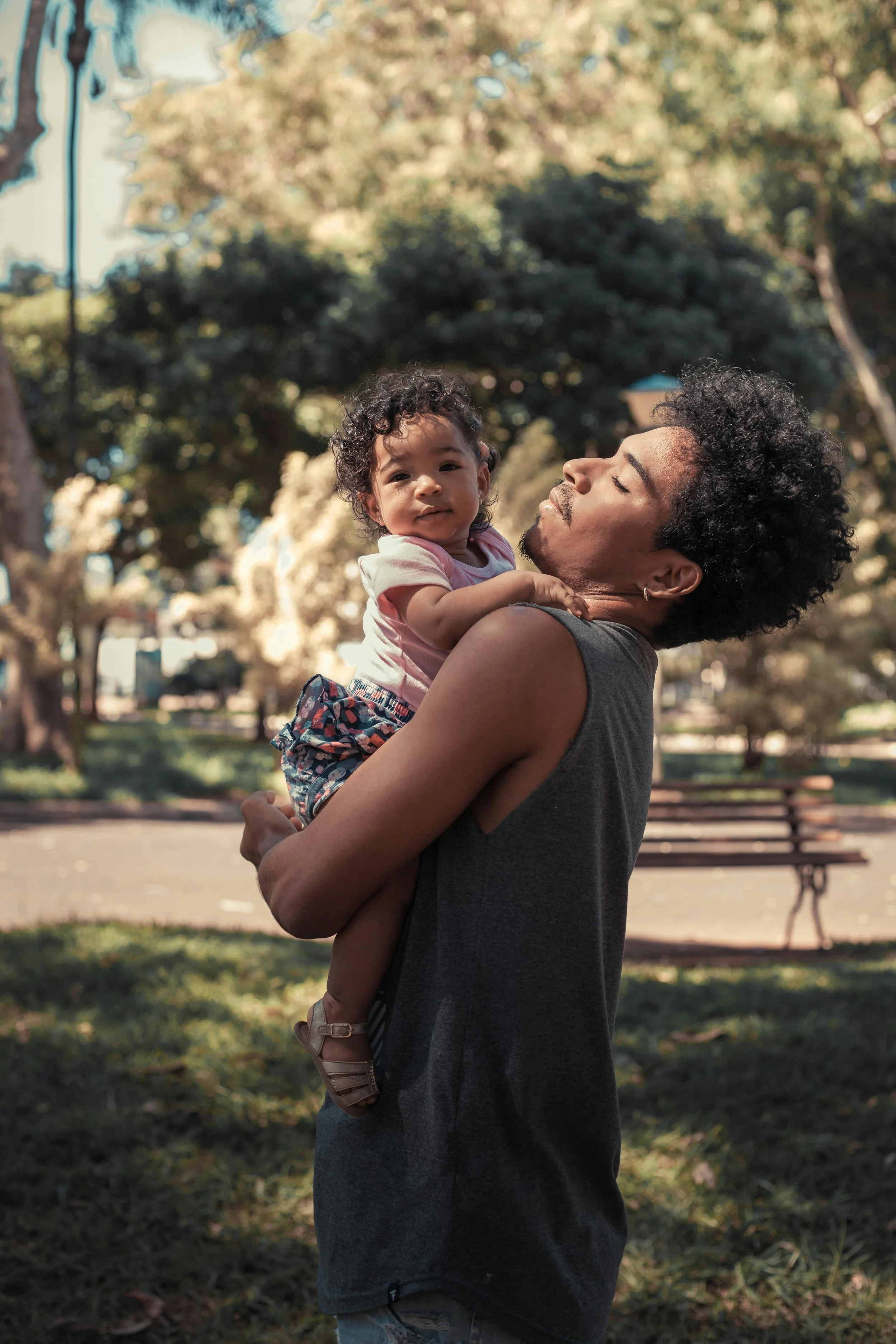A man holding a young girl in a park with trees and a bench in the background.