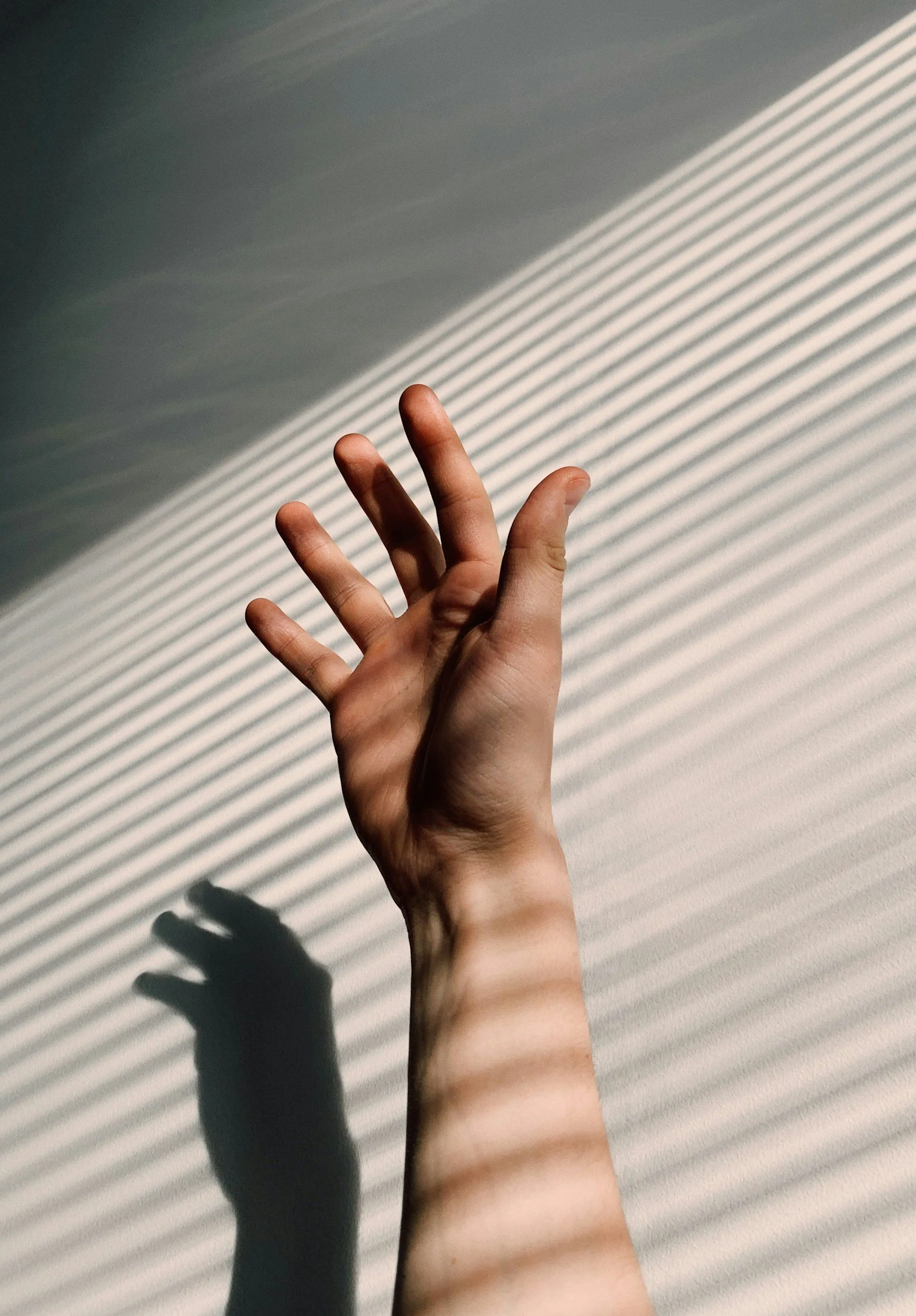 A human hand reaching upward, casting a shadow on a wall with diagonal striped light patterns.