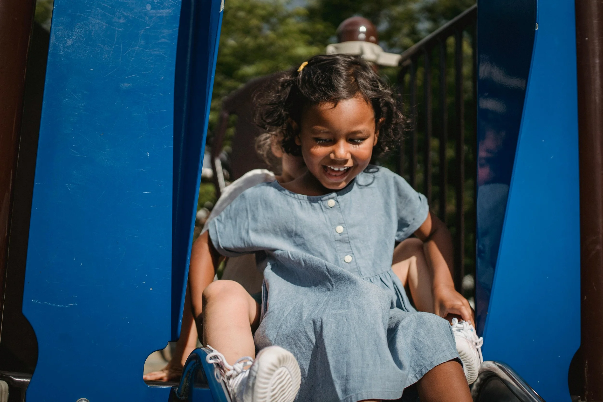 A young girl in a light blue dress sitting on a playground slide with a big smile, surrounded by playground equipment and greenery in the background.