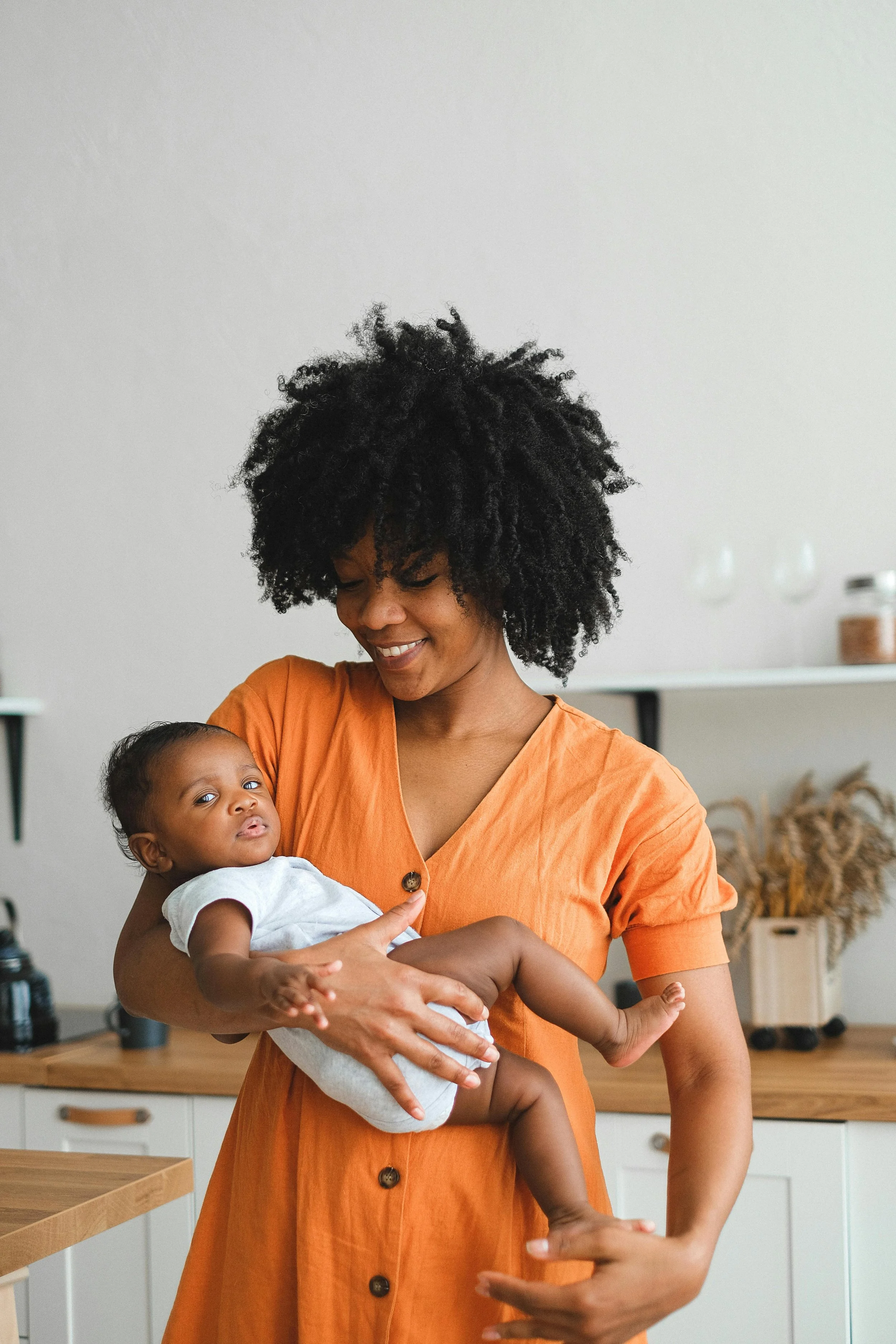 A woman with a big curly hairstyle holding a baby in her arms inside a kitchen with a smiling expression