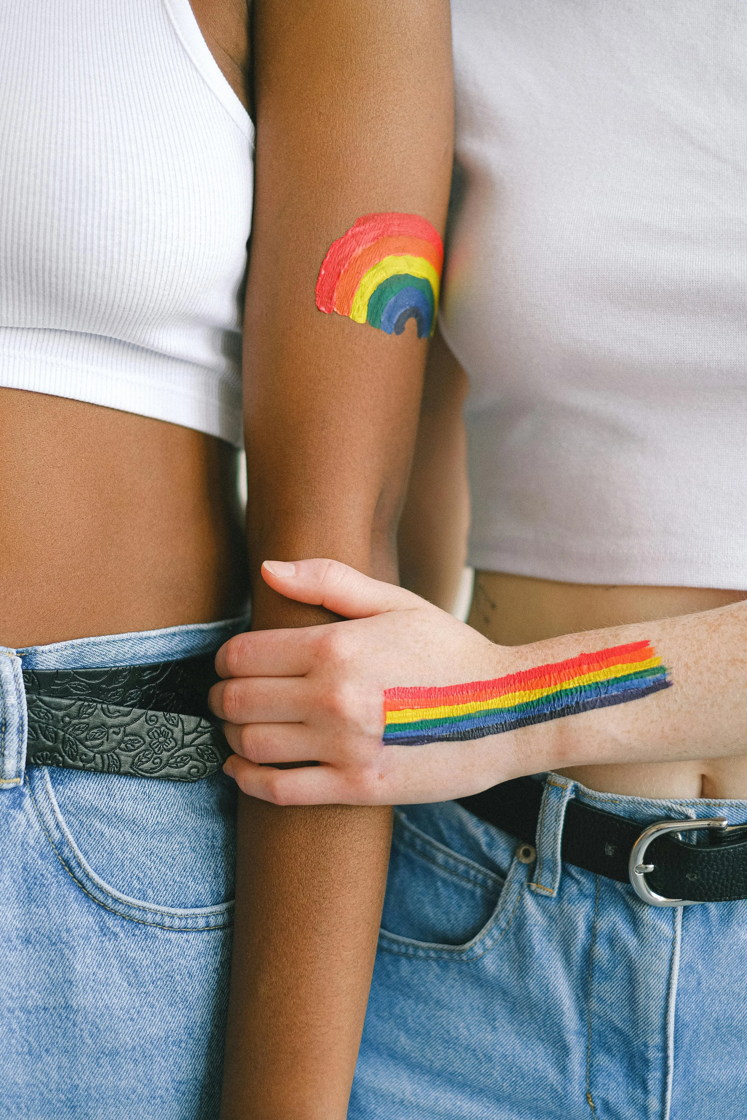 Close-up of two people with painted rainbow symbols on their skin, one with a rainbow on their arm and the other on their side, holding each other's arms, wearing casual clothing.