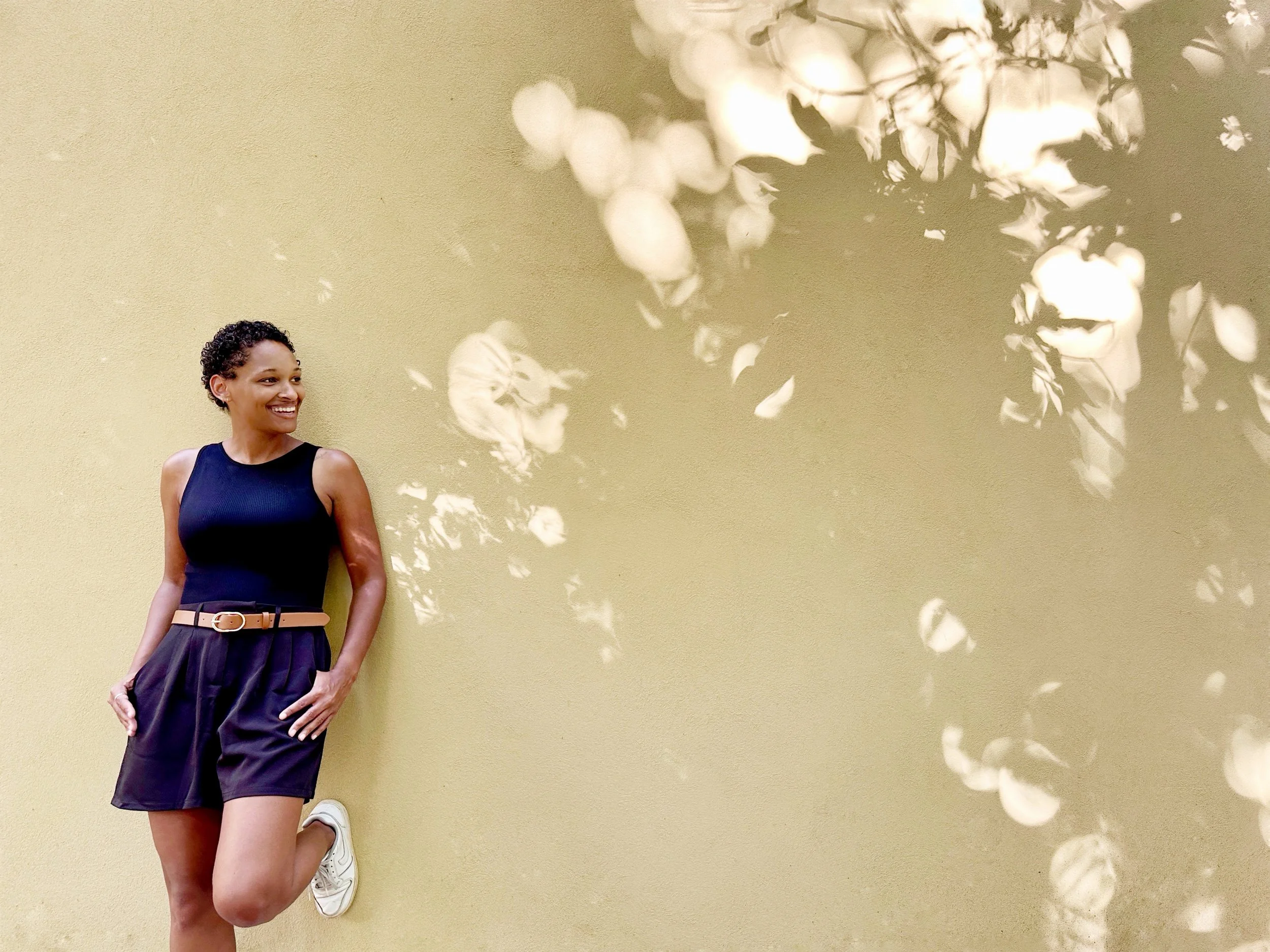 A woman in a black sleeveless top, dark shorts, and white sneakers leaning against a beige wall, smiling and looking to her left. Sunlight filter creates a pattern of bokeh and shadows on the wall and her face.