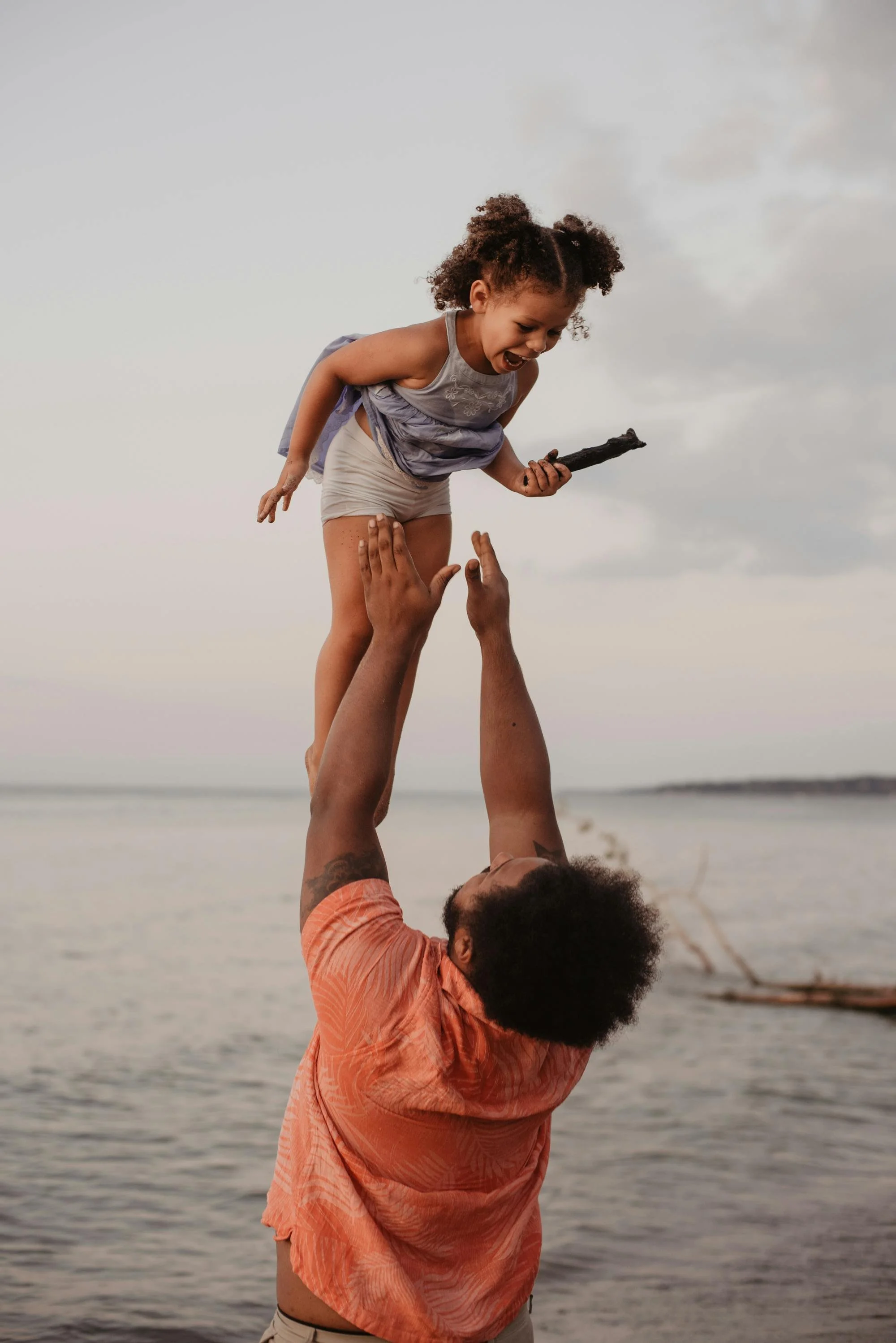 A man lifting a young girl in the air at the beach during sunset, both appearing joyful.