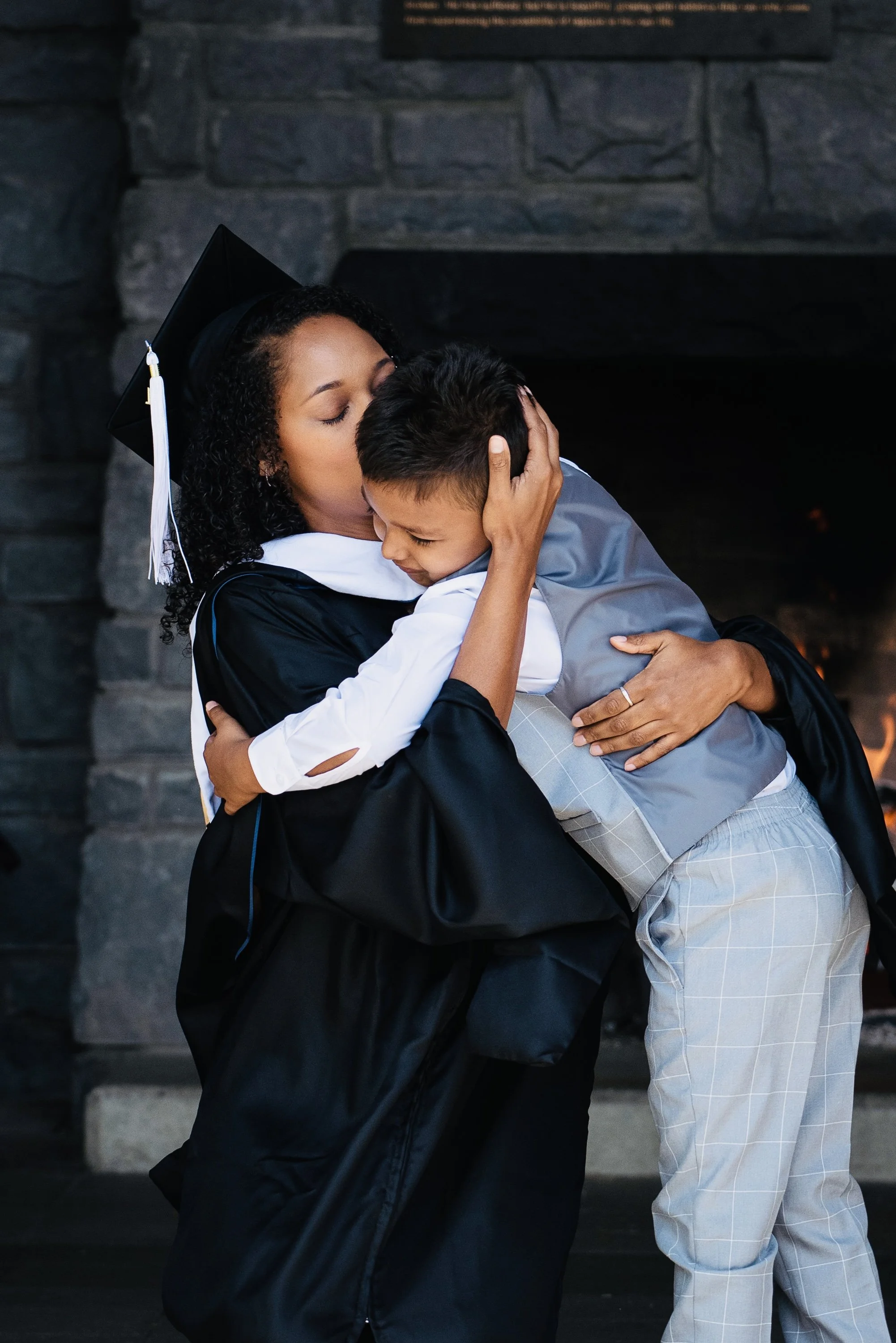 A woman in graduation attire hugging and kissing a young boy in front of a fireplace.