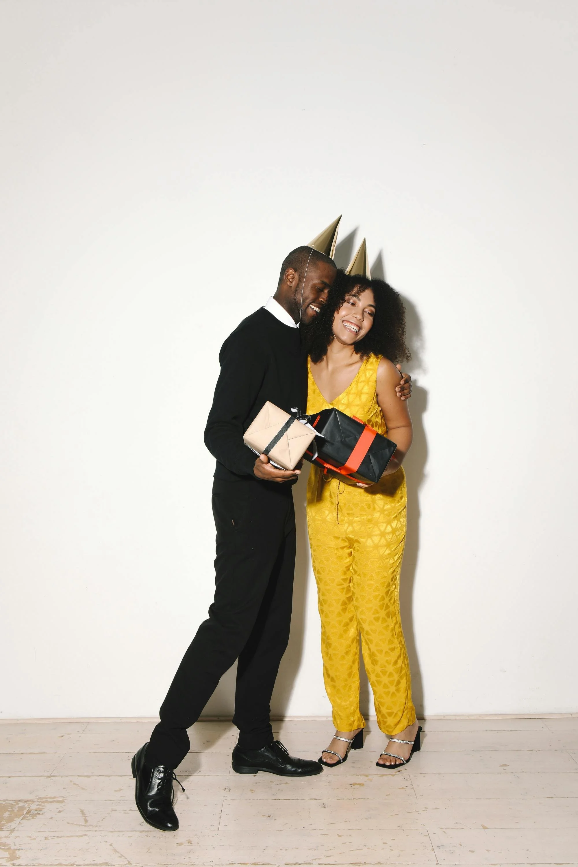A man and woman celebrating with party hats, holding gifts, smiling, standing against a plain white wall, on a wooden floor.