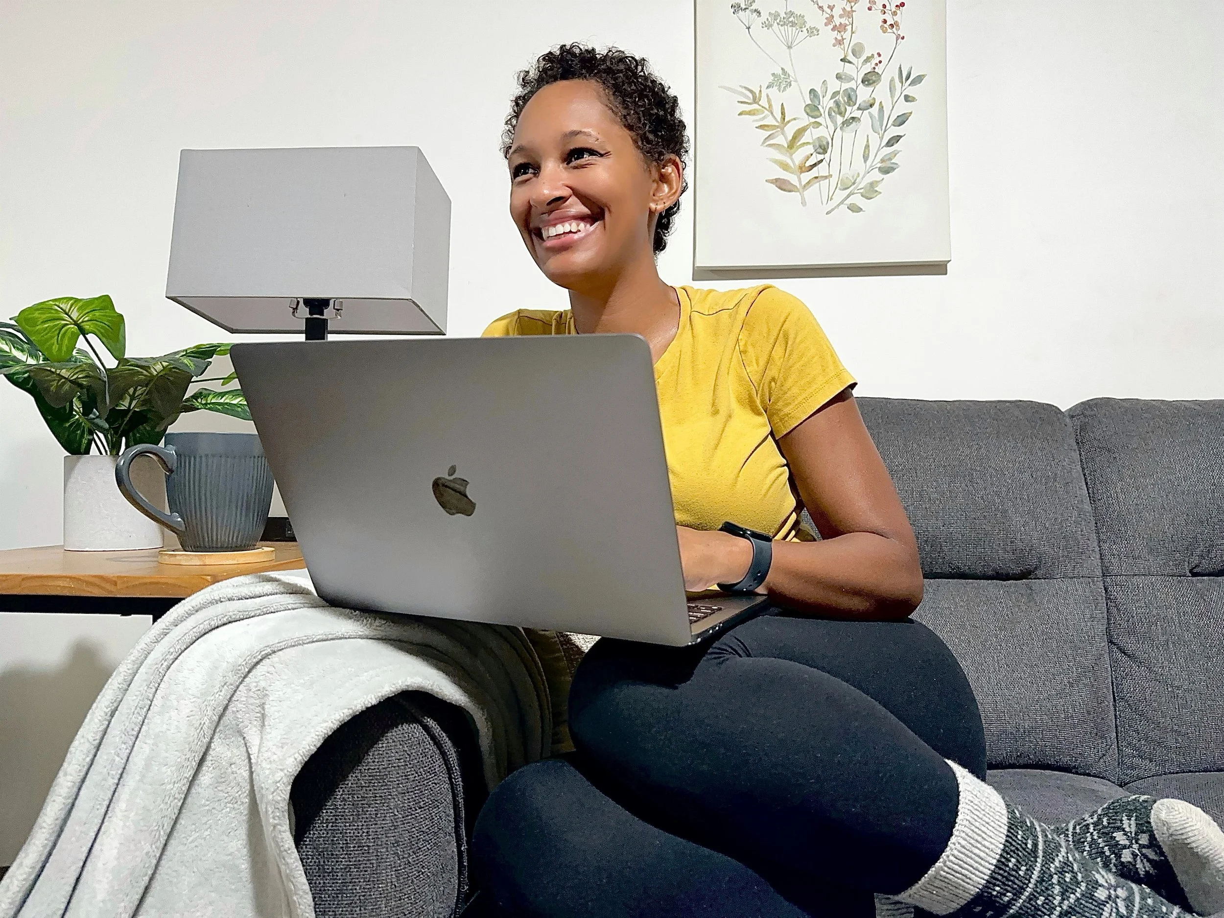 A smiling woman with short curly hair, wearing a yellow t-shirt, sitting on a gray couch with a laptop on her lap, surrounded by houseplants and a lamp on a side table.