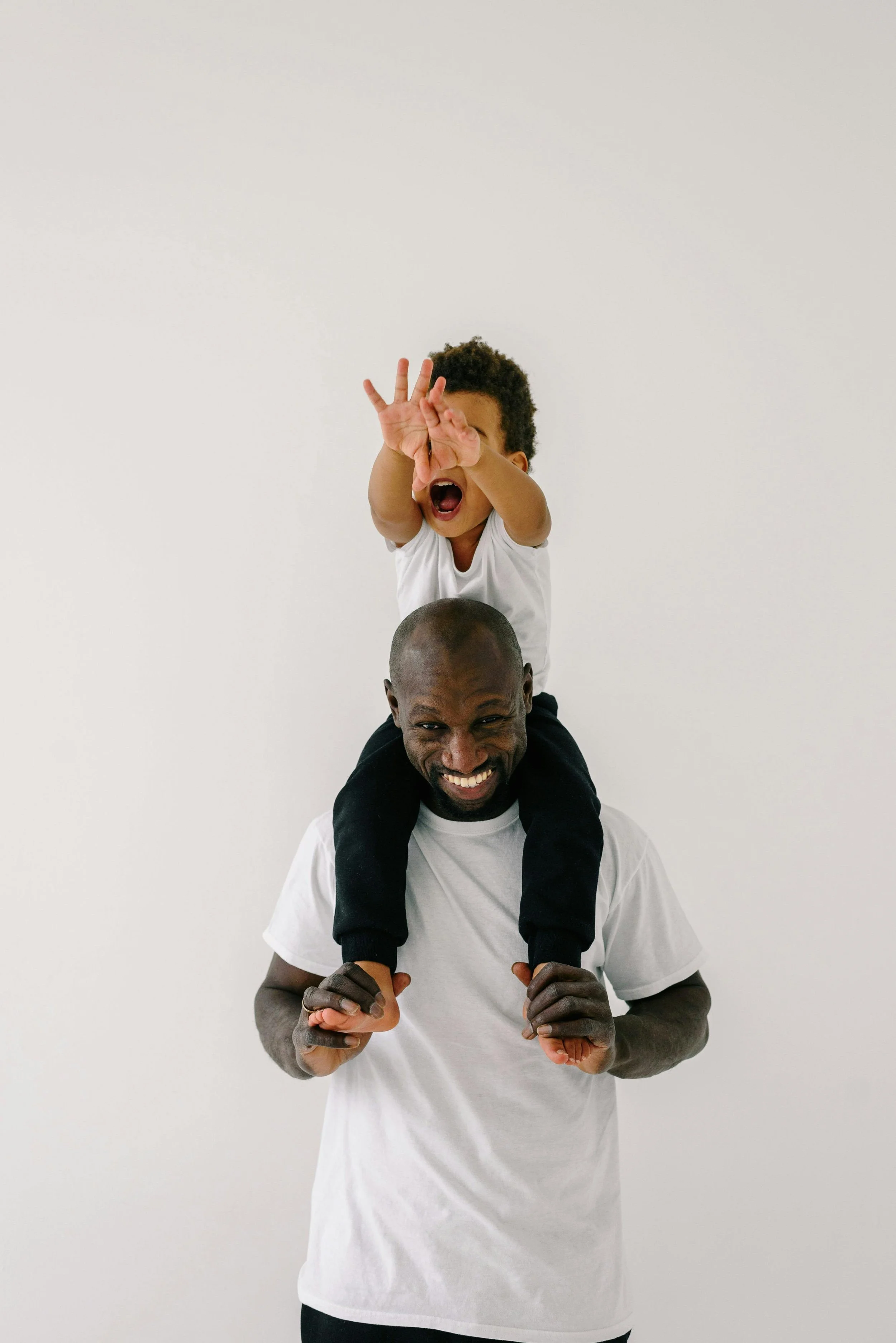 A man carrying a young boy on his shoulders, both smiling. The boy is reaching out with his hand, and the background is plain white.