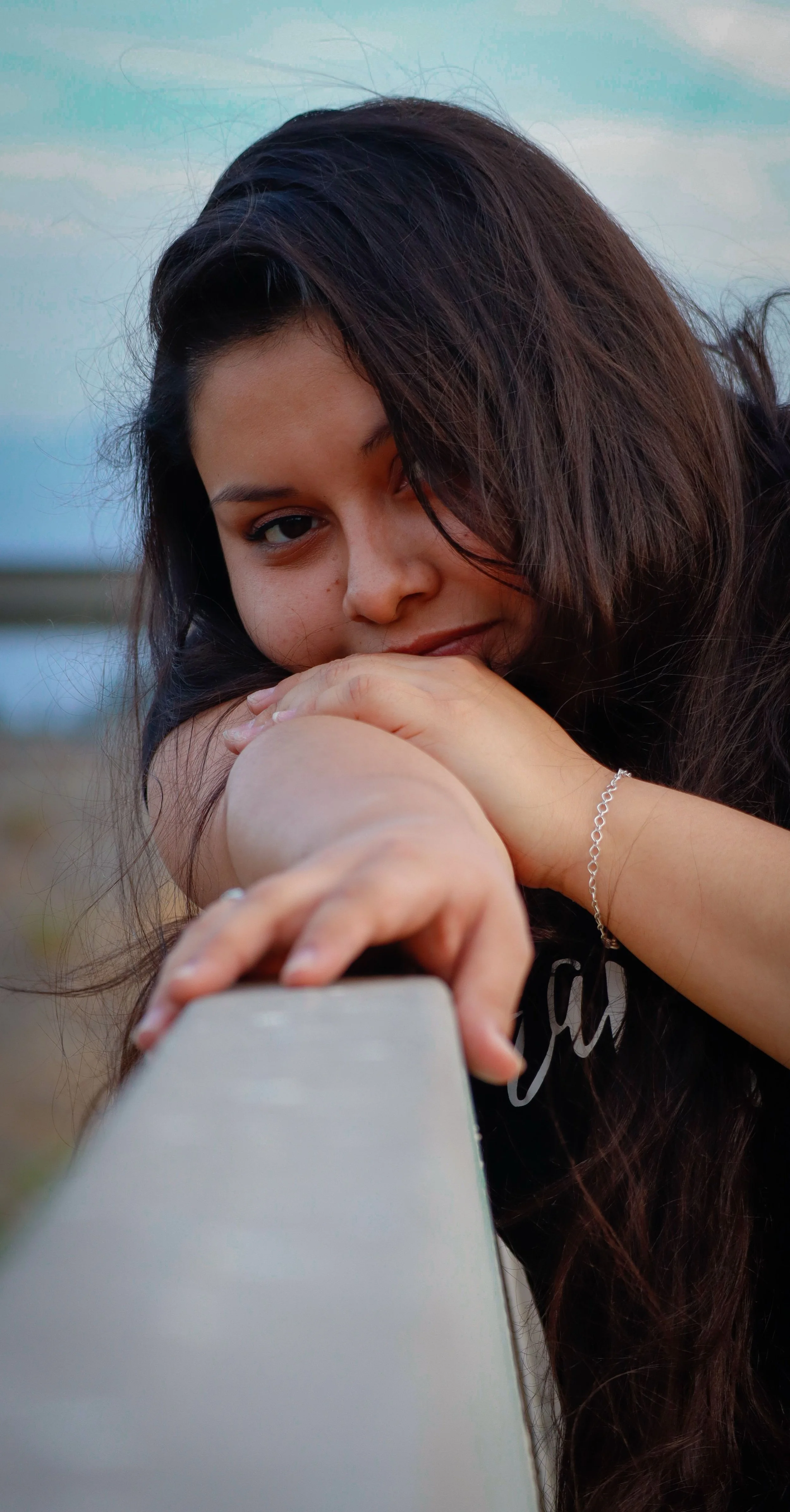 Woman resting her chin on her folded arms, looking thoughtfully toward the camera outdoors.”