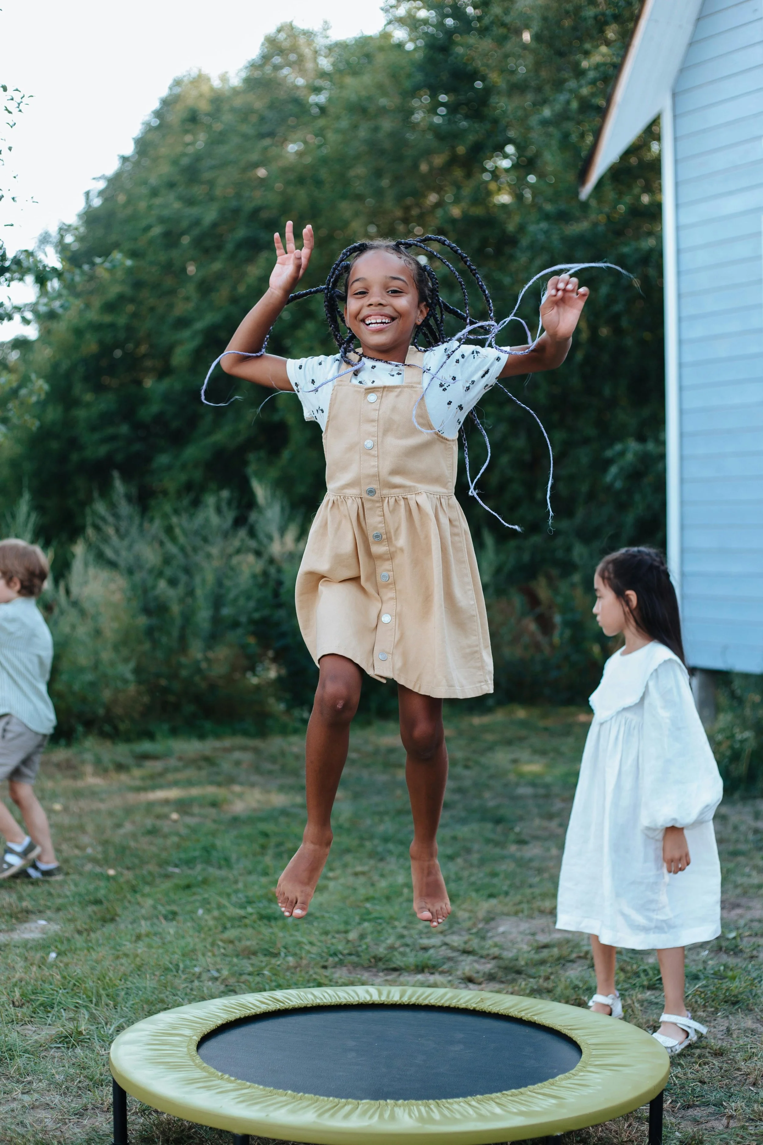 A young girl joyfully jumping on a mini trampoline outdoors with other children nearby, surrounded by greenery.