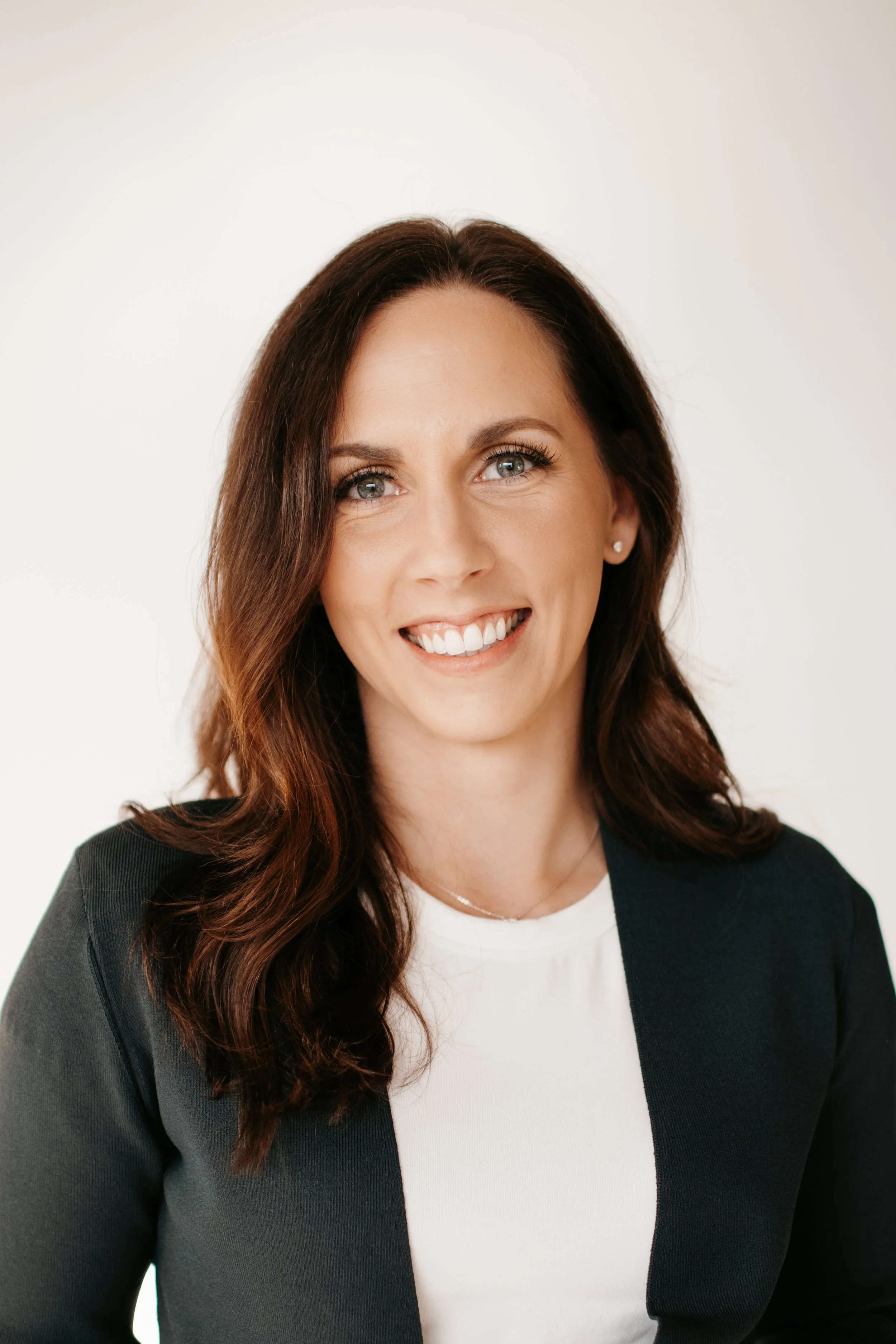 A woman with long brown wavy hair, blue eyes, and light makeup smiling for a portrait against a plain white background, dressed in a white top and black blazer.