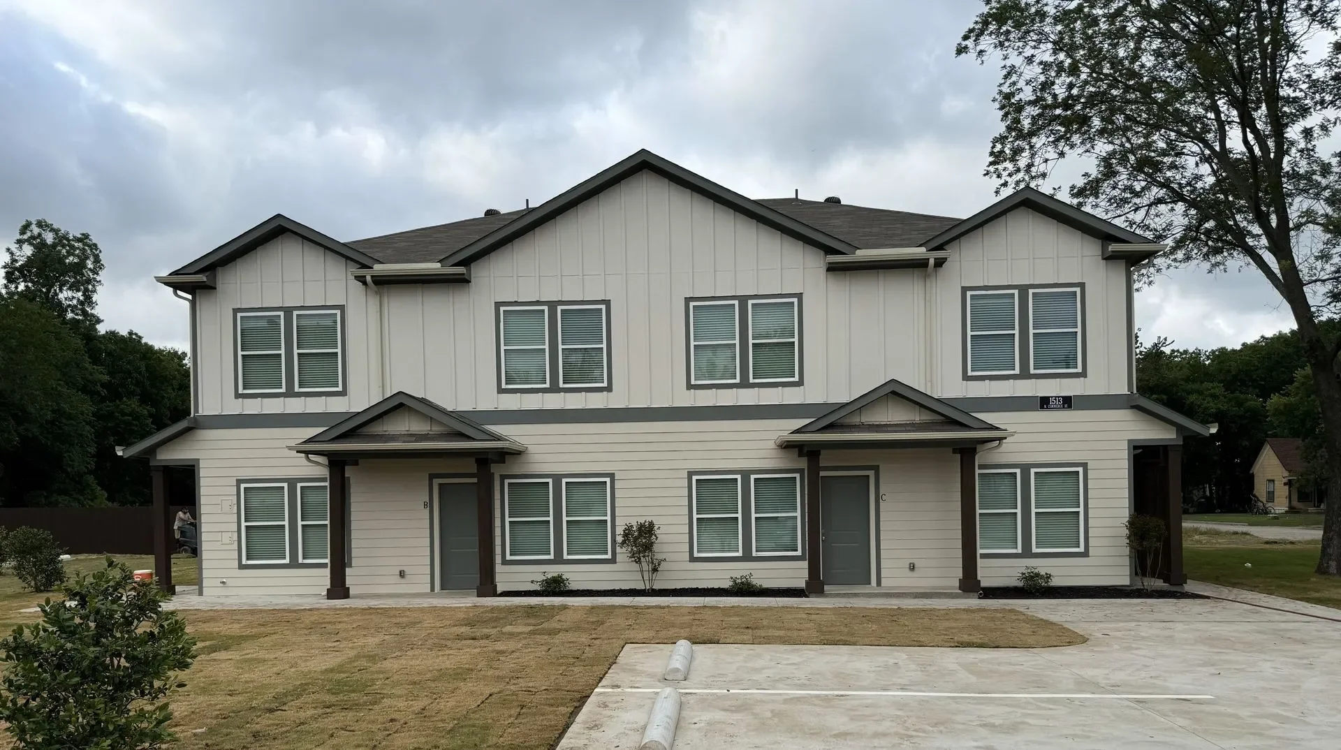 A newly built two-story apartment complex with beige siding and dark brown trim. The building has multiple windows with white trim, and two entrances with small covered porches. There is a concrete parking lot in front and a grassy area with small shrubs.