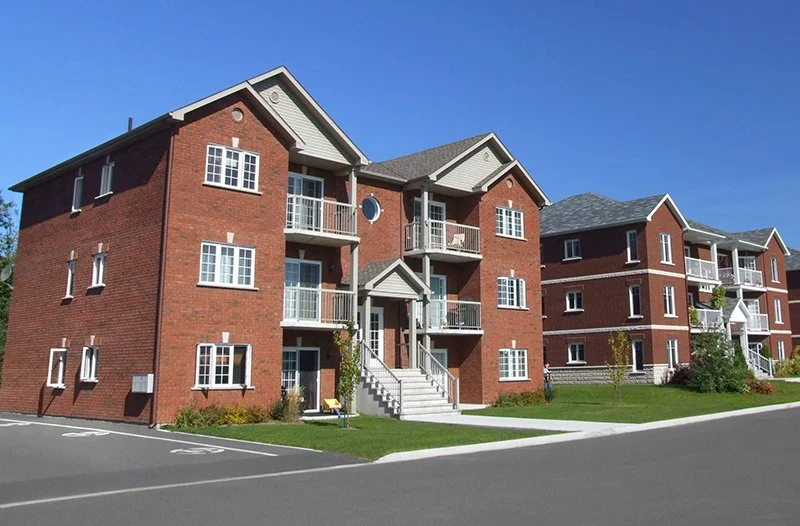A three-story red brick apartment building with white trim and balconies, situated on a well-maintained lawn with a sidewalk and parking lot, under a clear blue sky.