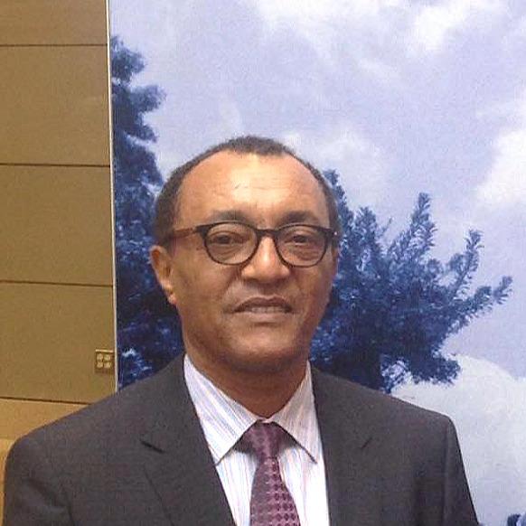 A middle-aged man with glasses and short hair, wearing a dark suit, white striped shirt, and purple tie, standing in front of a cloudy sky background.