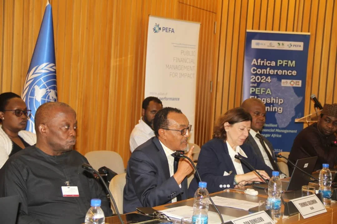 Panel of six people seated at a conference table with microphones, with African-themed banners and a United Nations flag in the background, during the Africa PFM Conference 2024.