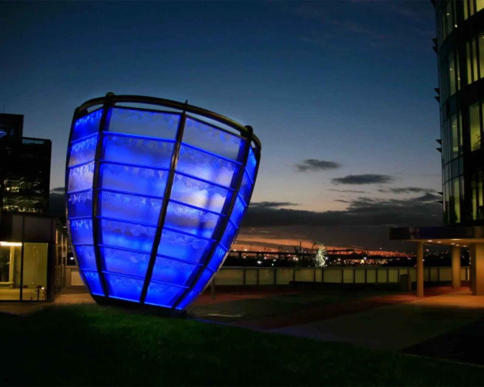 A modern building with a large, illuminated blue glass structure on a rooftop terrace during dusk, with a city skyline and fireworks in the background.