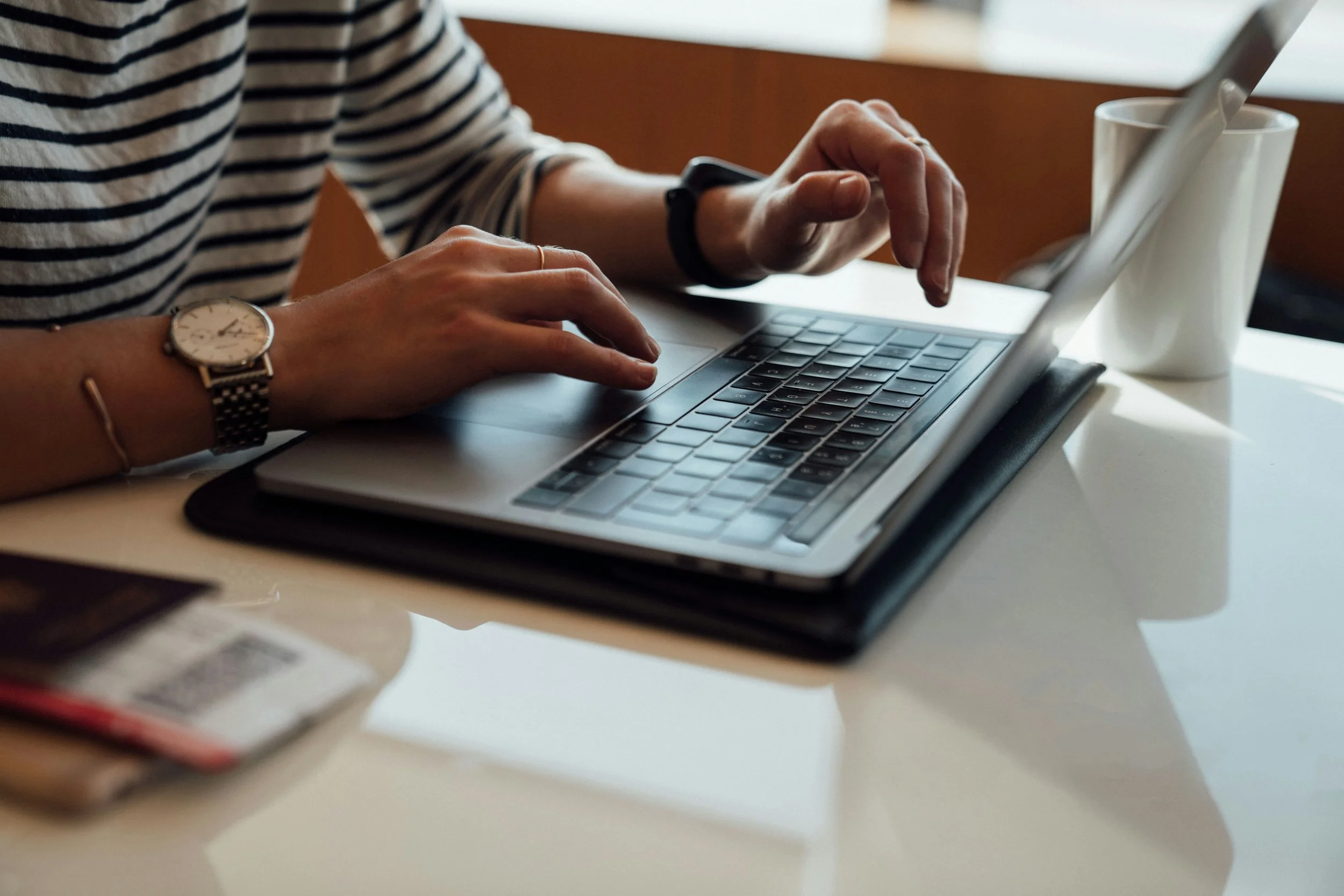 Person typing on a laptop with a striped shirt, wristwatch, and phone nearby on a white table.