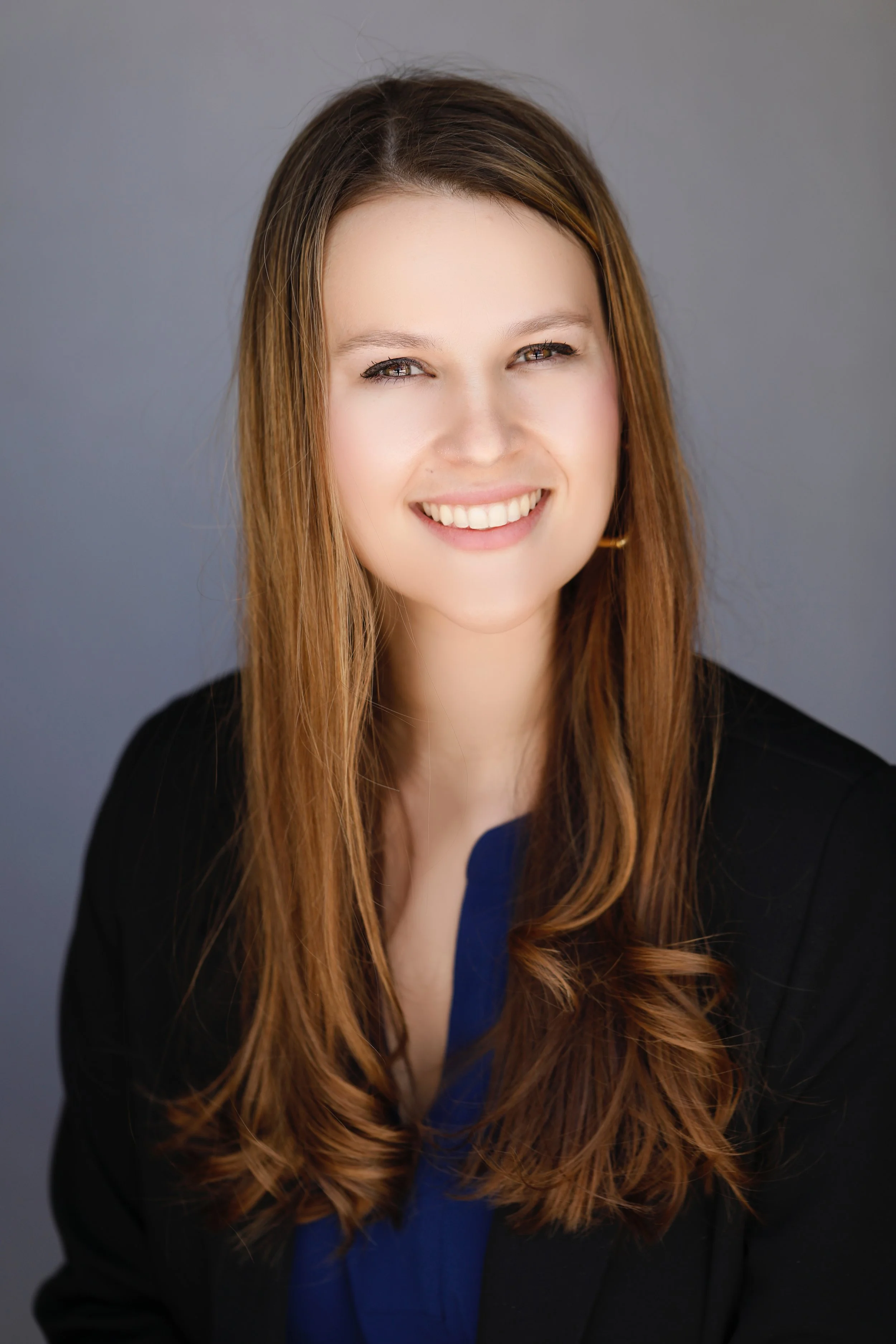 Headshot of a young woman with long wavy hair, smiling, wearing a dark blazer and a blue top against a gray background.