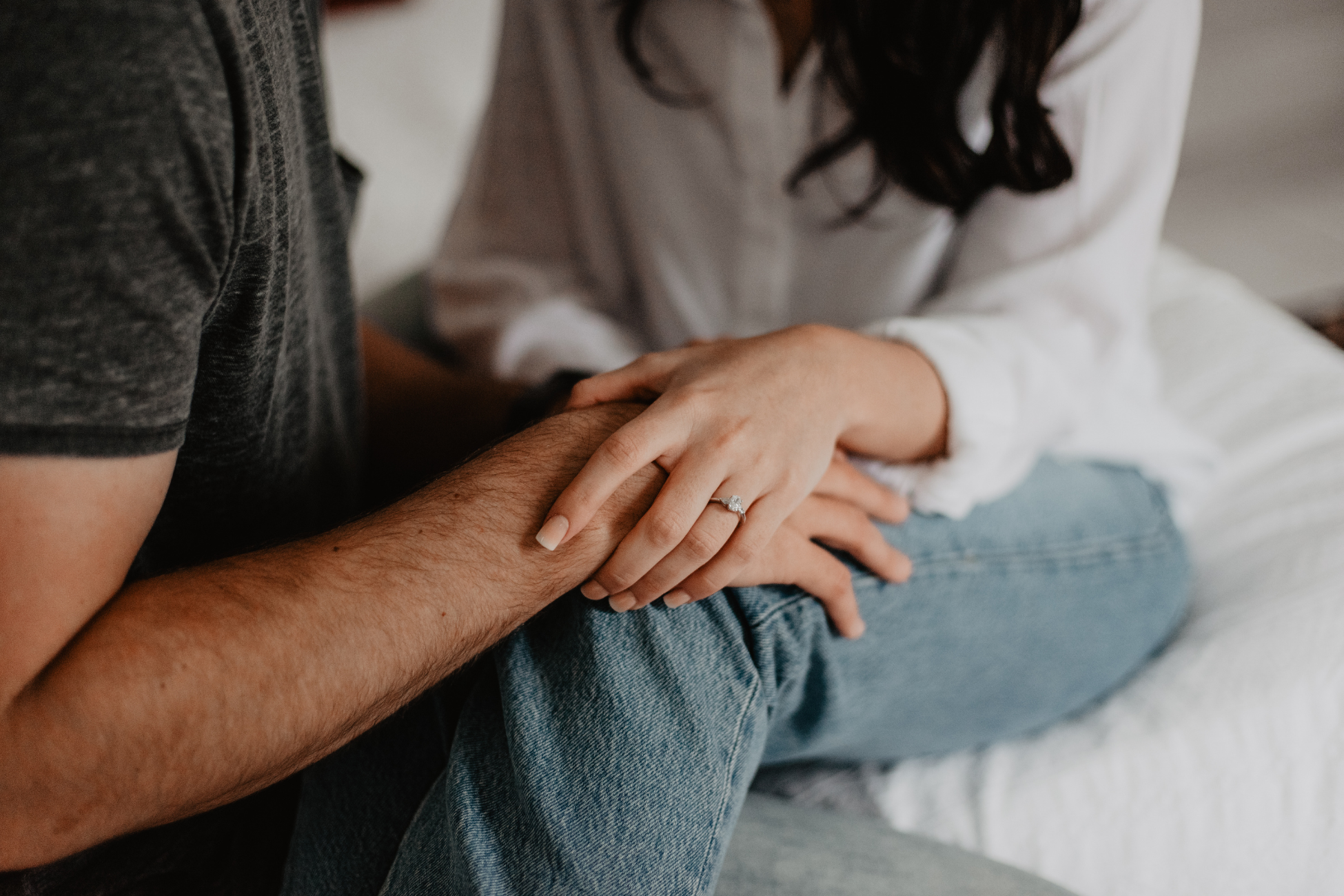 A woman wearing a white shirt is holding a man's hand which has an engagement ring on her finger.