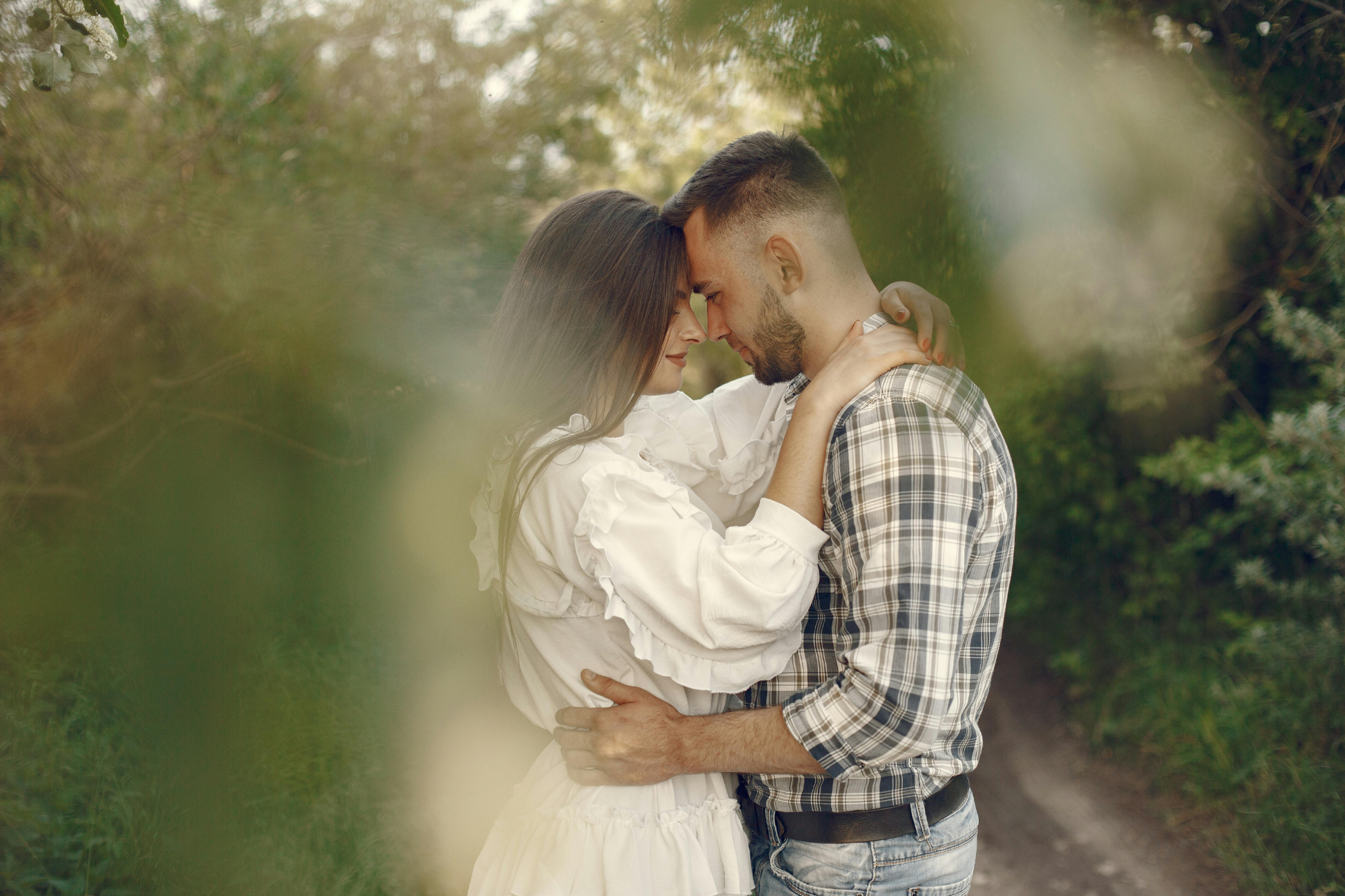 A man and woman are close together outdoors, touching foreheads and holding each other tenderly, surrounded by greenery.