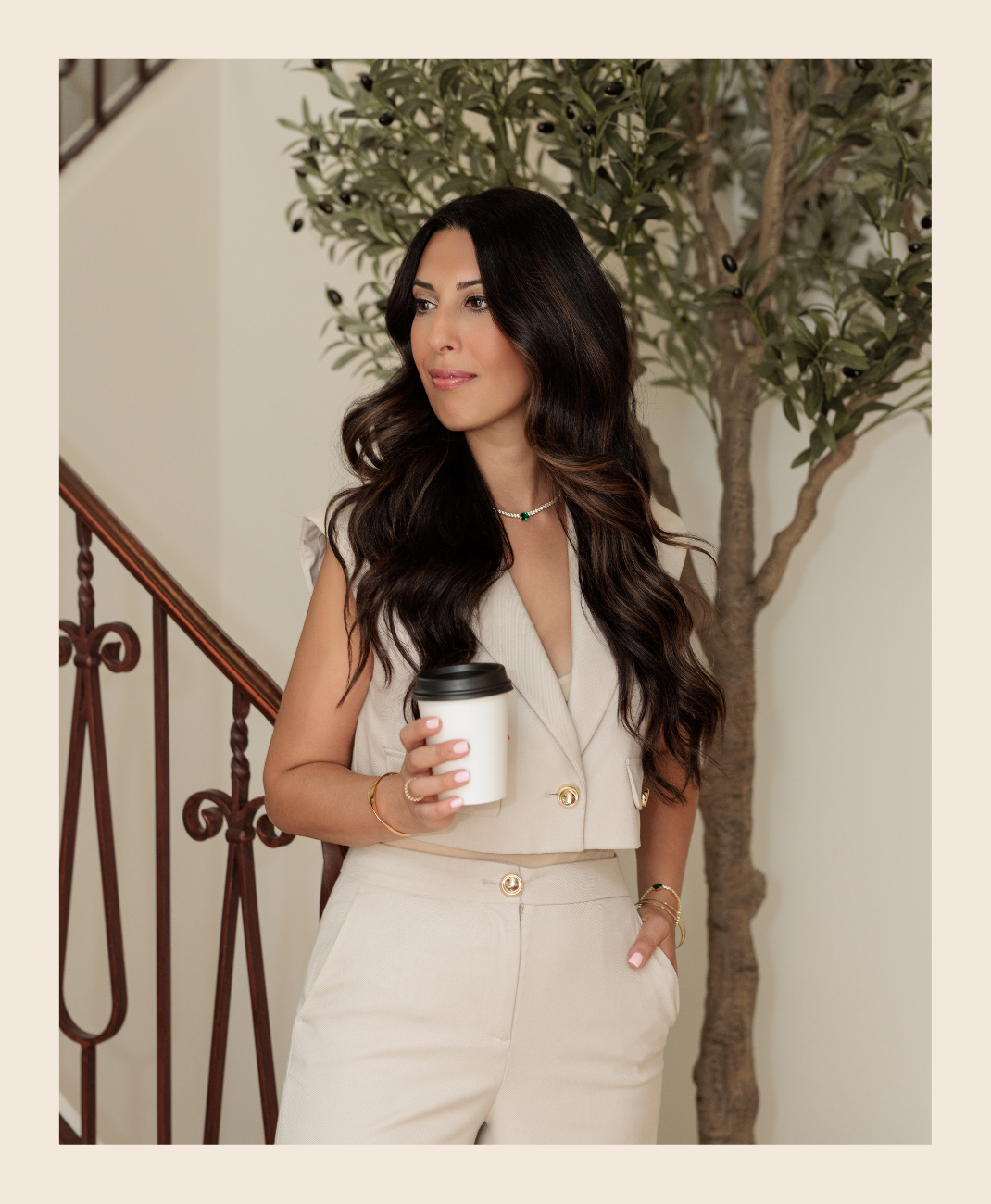 Woman with long dark wavy hair wearing a beige sleeveless blazer and pants holding a coffee cup, standing near a staircase and a green potted plant.