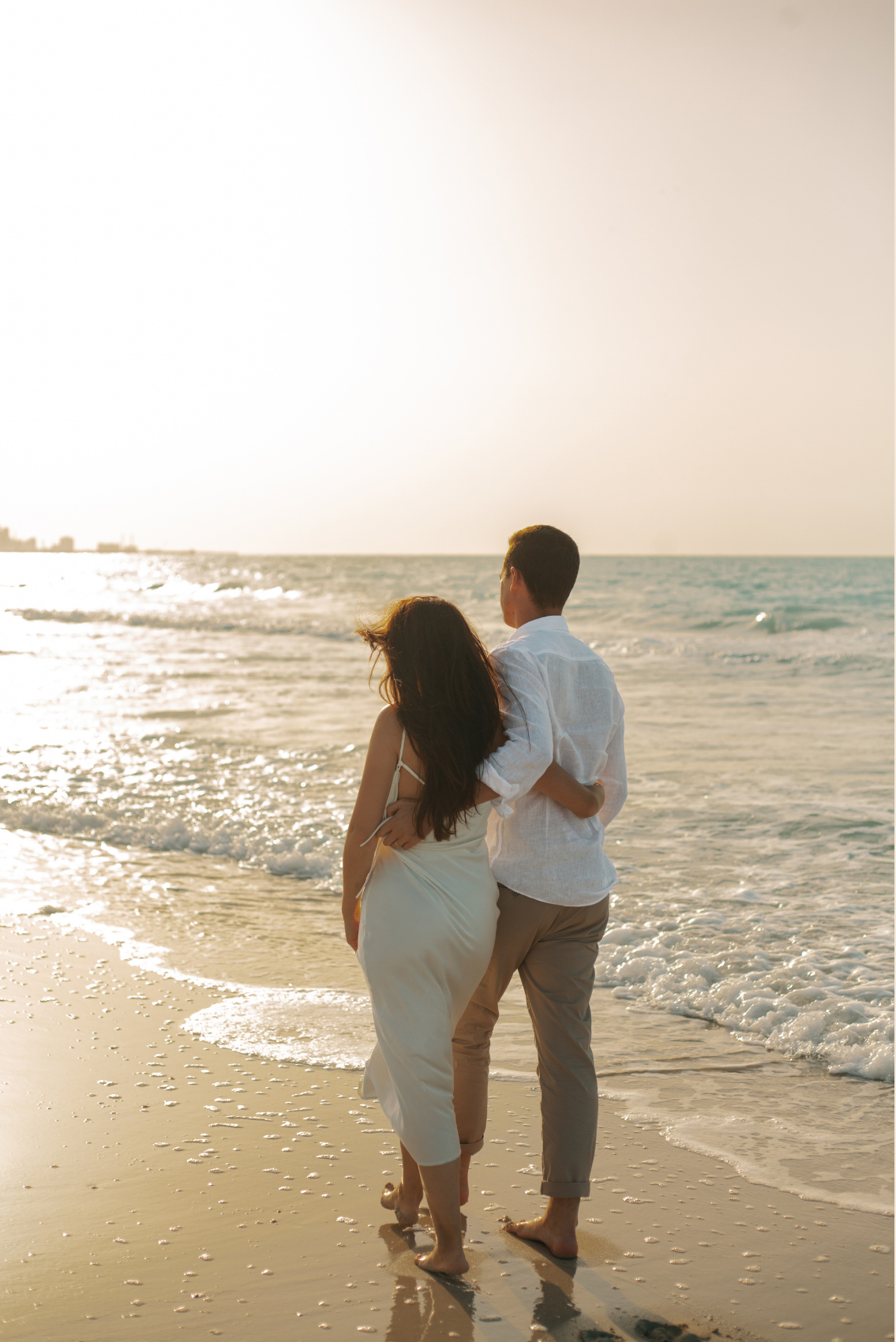 A couple walking along the beach at sunset, with the ocean waves near their feet.