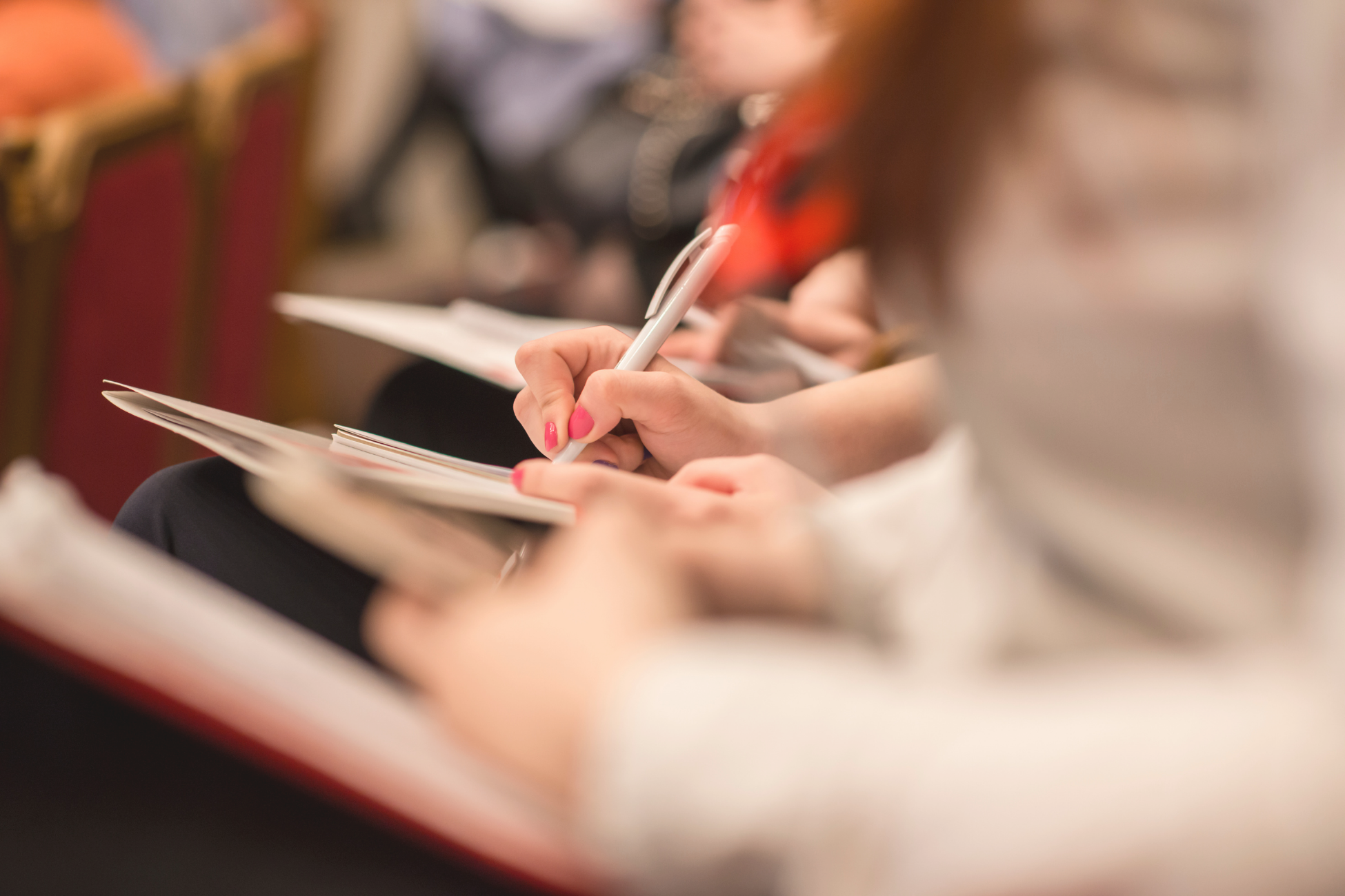 People seated in a conference or lecture, taking notes with pens and notebooks.