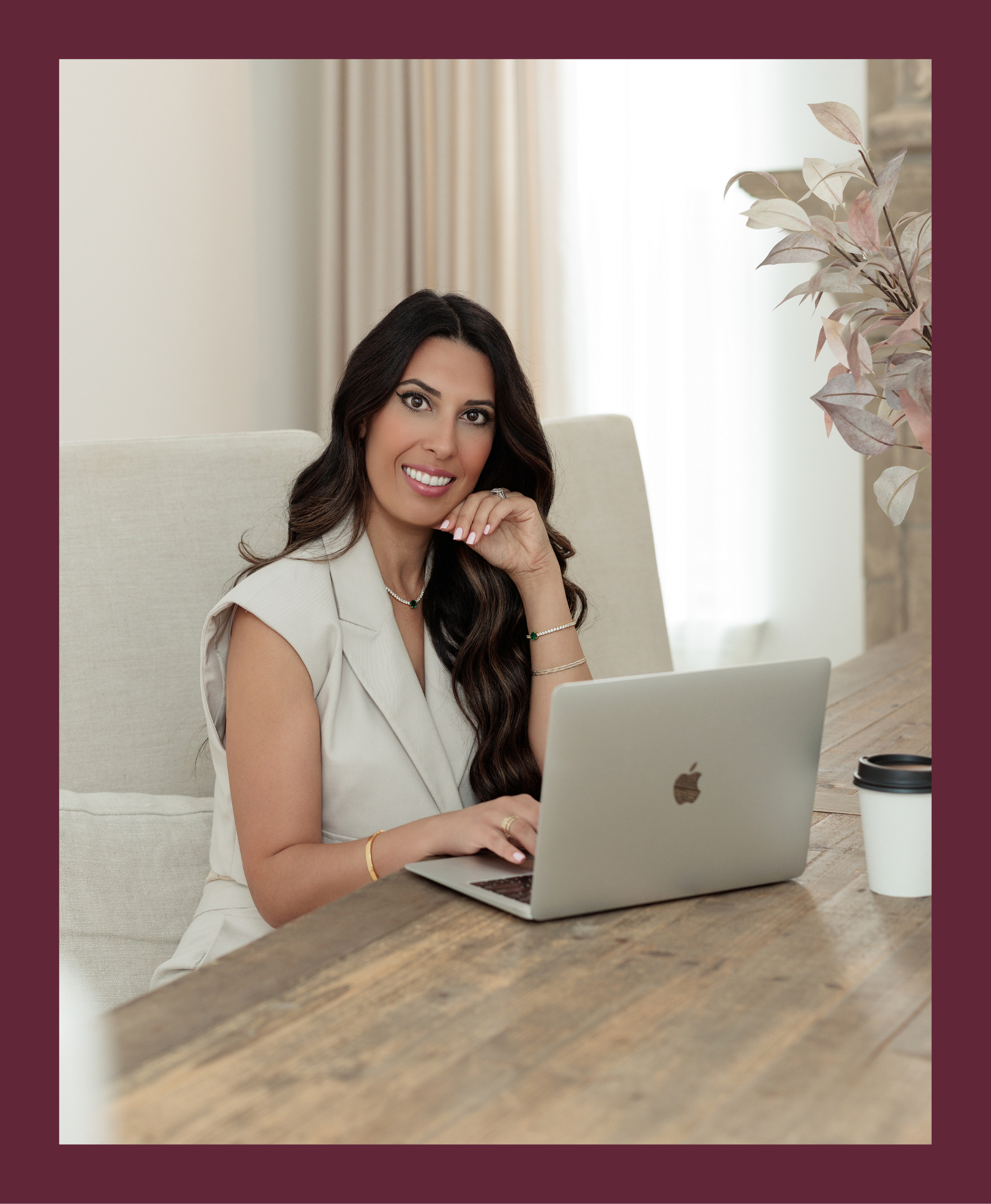 A woman with long dark hair sitting at a wooden table using a silver MacBook laptop, smiling, with a coffee cup nearby, in a bright room with cream curtains and neutral decor.